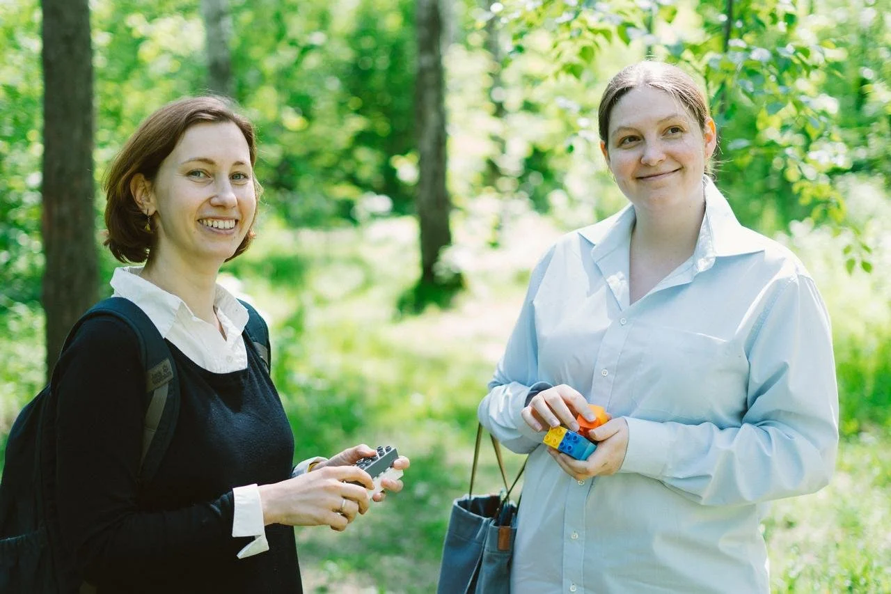 Two women smiling outdoors in a green, wooded area, holding toy tools, with one carrying a backpack.