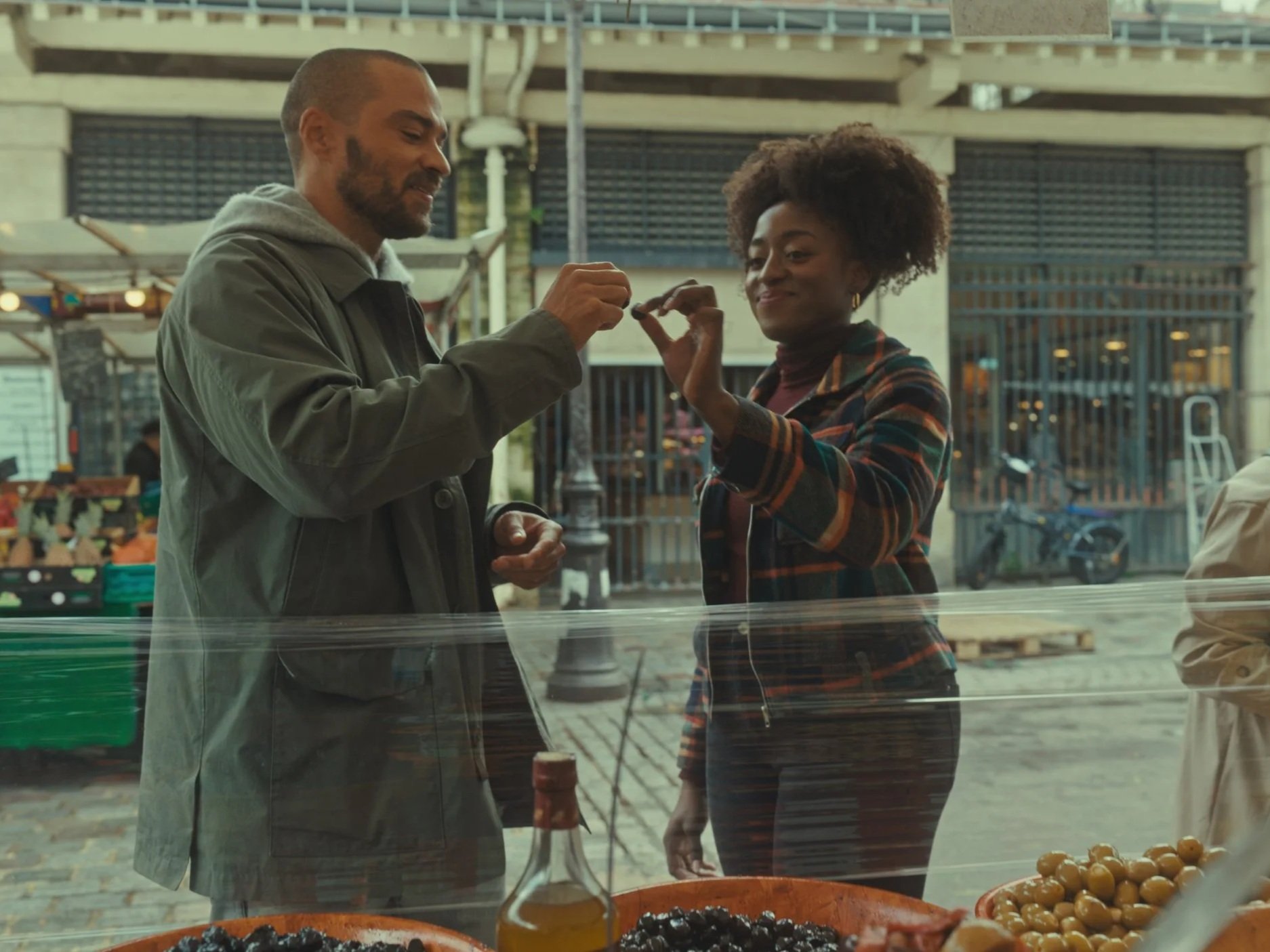 A man and woman at an outdoor market, holding a small object and smiling. The market stall has jars of olives and a bottle of olive oil in the foreground.