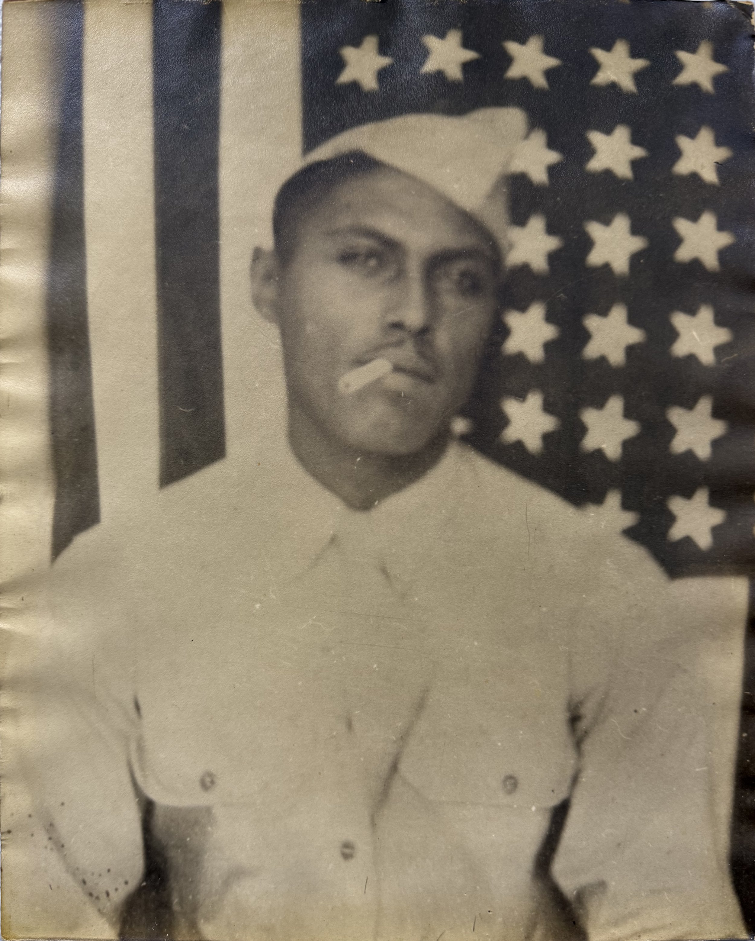 A black-and-white photograph of a young man in a military uniform, posed in front of an American flag background, with a cigarette in his mouth.