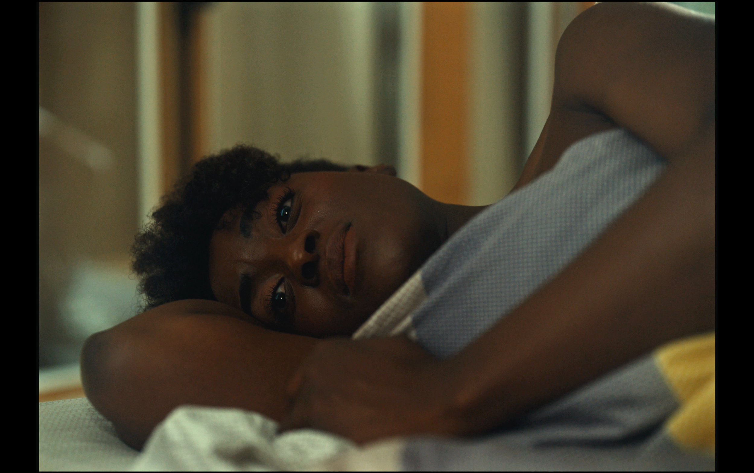 A woman lies on a bed, facing the camera with her head resting on her arm, featuring a thoughtful or pensive expression.