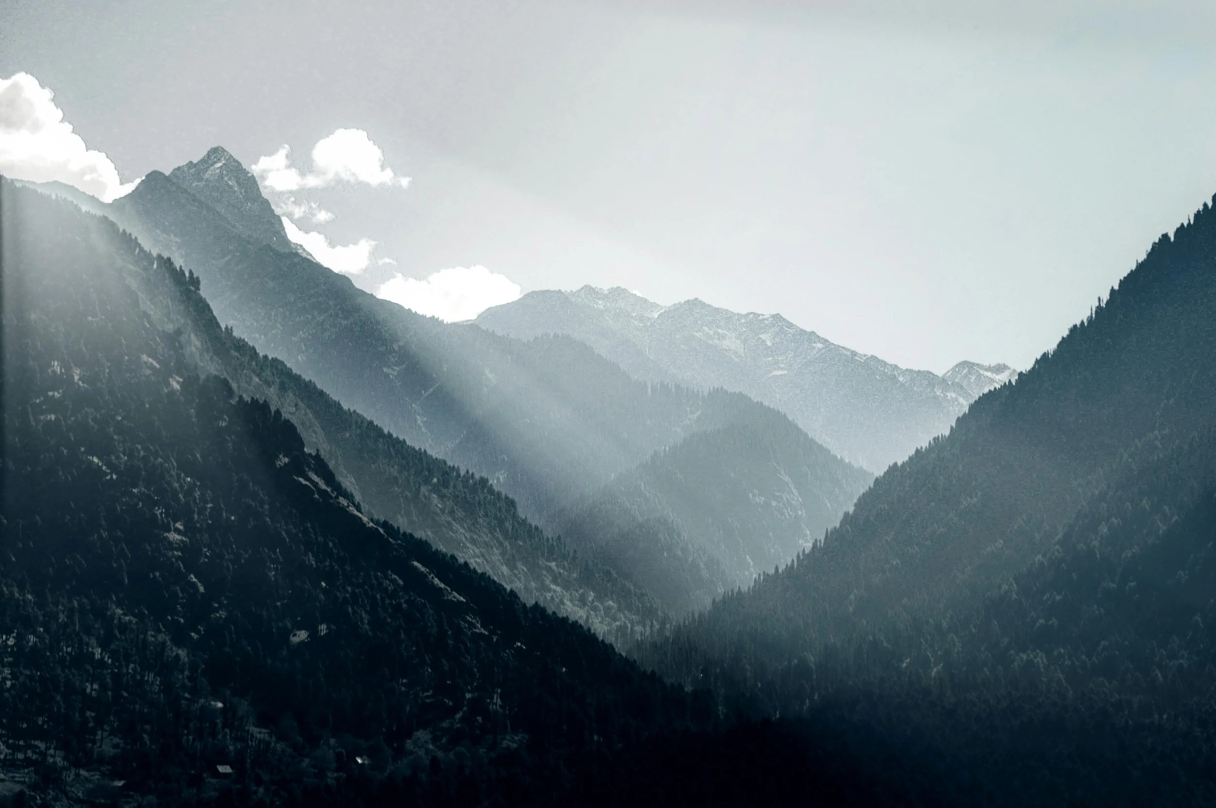 Mountain range with multiple peaks, covered in trees, with sunlight shining over the mountains and some clouds in the sky.