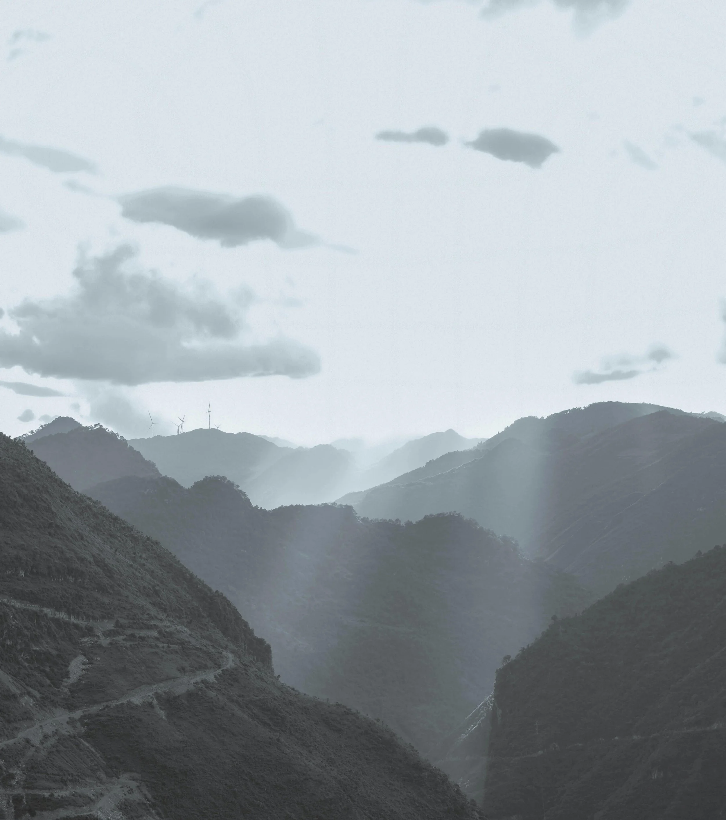 Photo of a mountain landscape with multiple ridges and valleys, cloudy sky, and wind turbines on distant peaks.