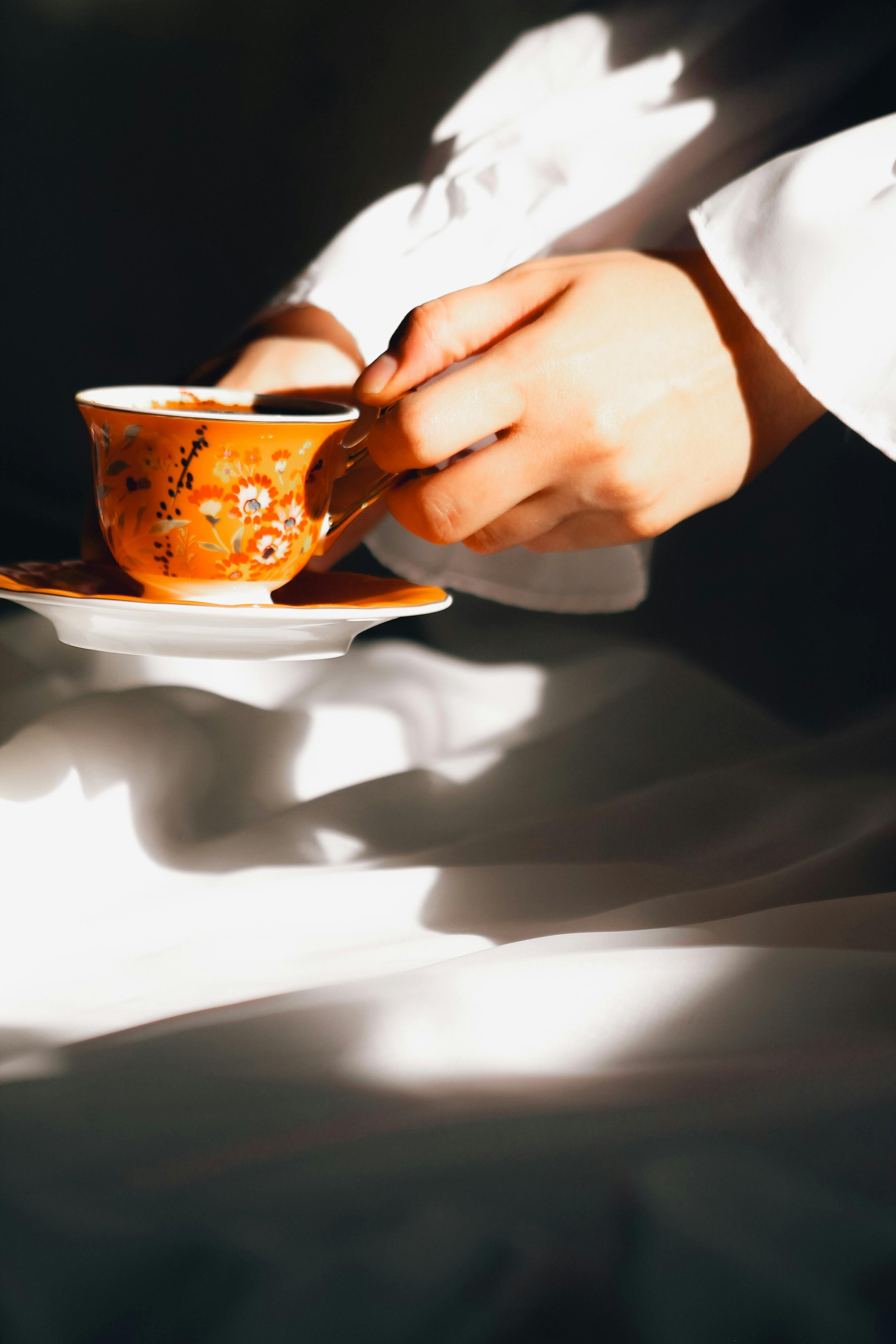A person's hand holding a floral-patterned tea cup over a matching saucer, with a dark liquid inside, on a white surface with shadows.