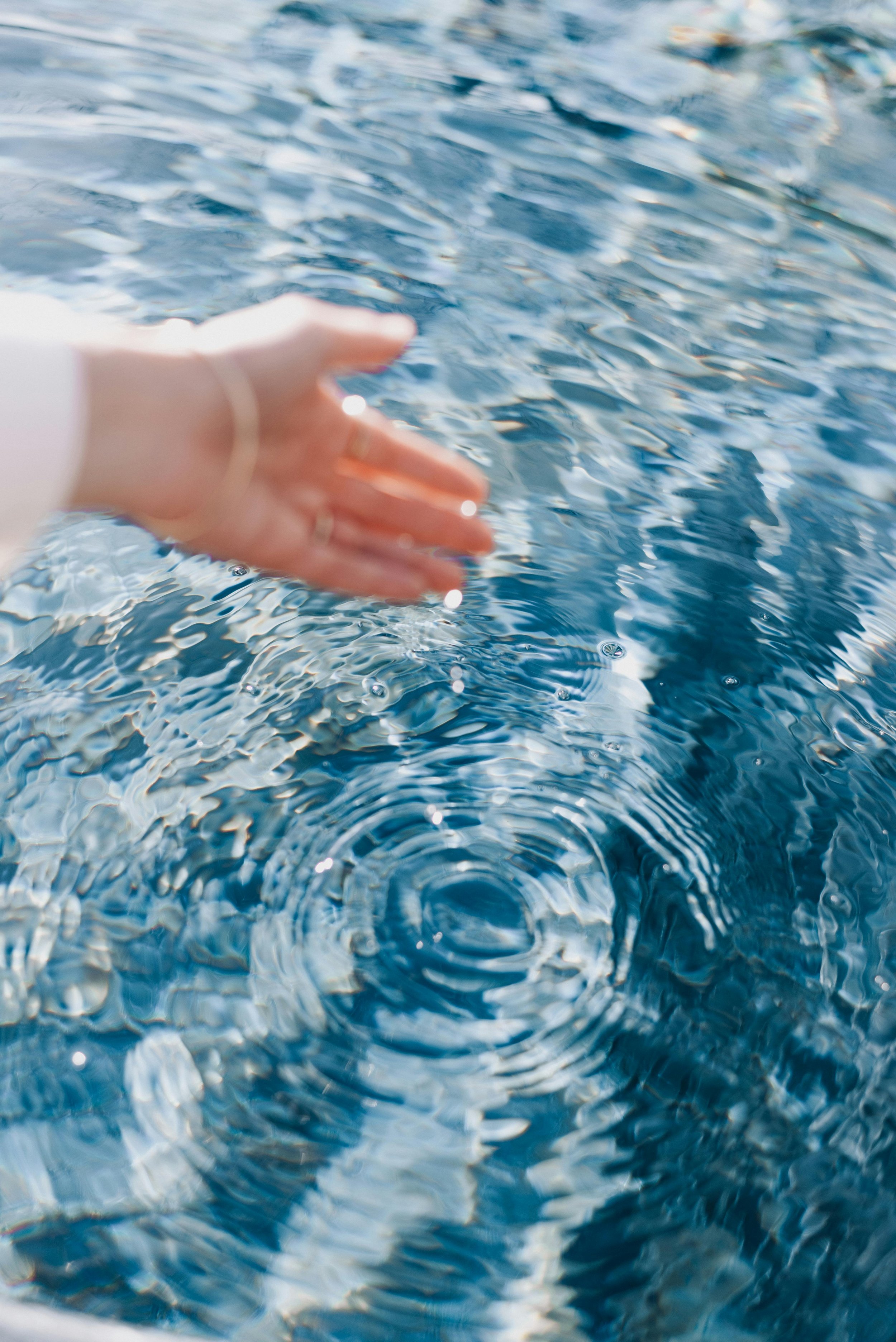 A hand with a gold bracelet reaching towards the surface of a body of water, creating ripples.
