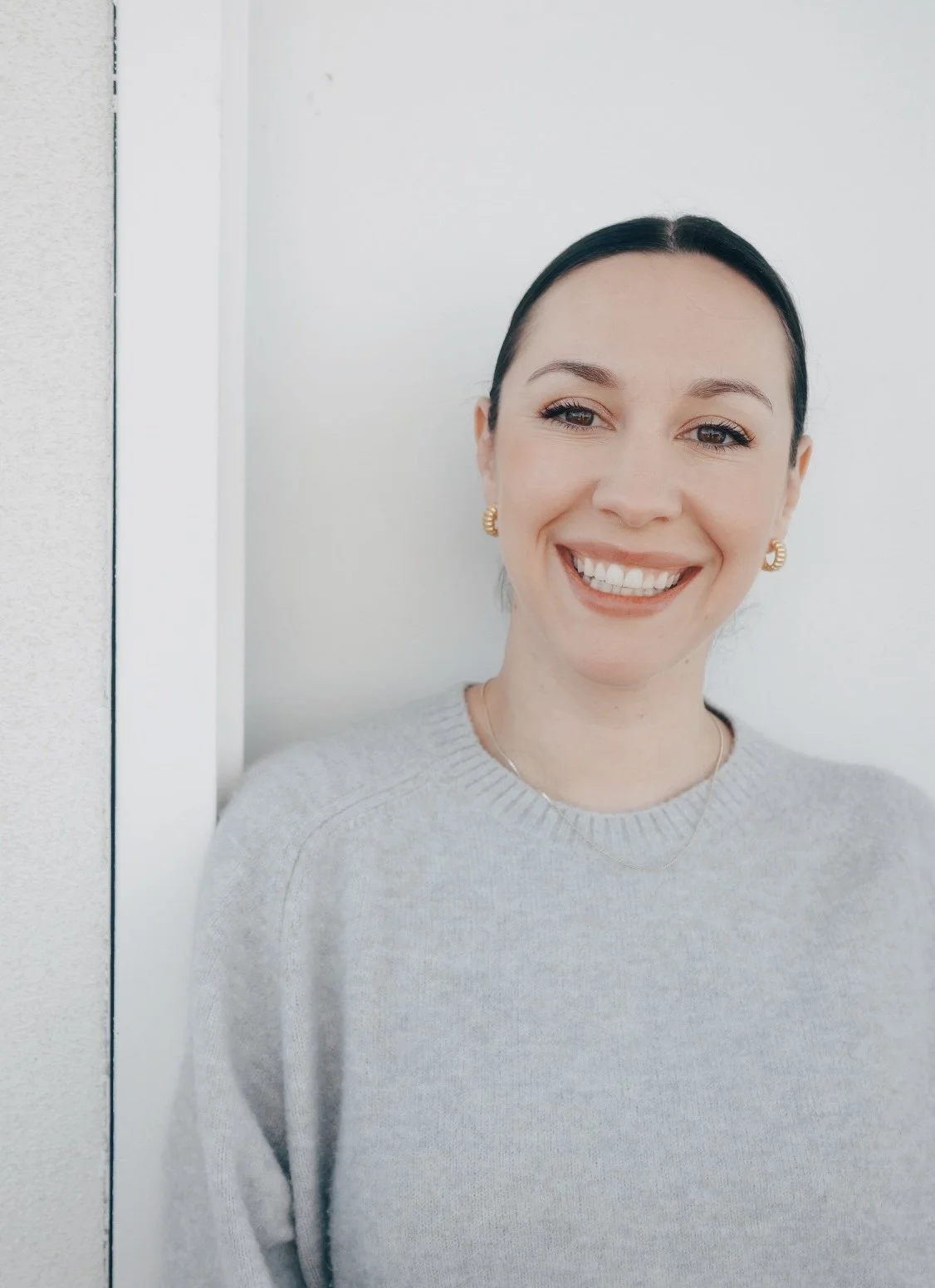 A woman smiling with dark hair pulled back, wearing gold earrings and a light gray sweater, standing next to a white wall.