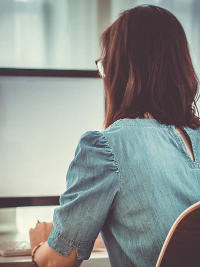 A woman with shoulder-length brown hair working at a desk in front of a computer, seen from behind.