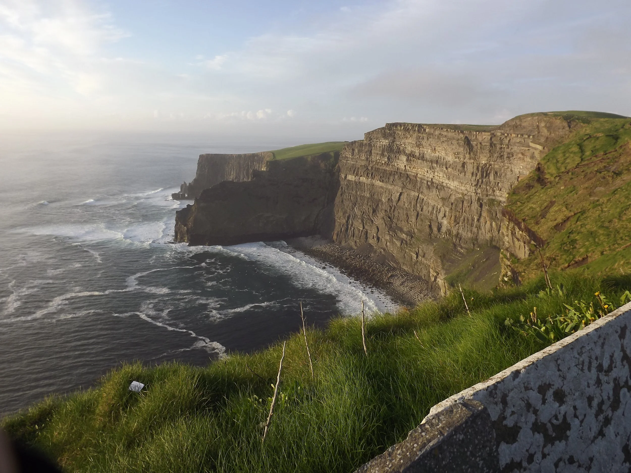 Cliffs along a rugged coastline with green grass in the foreground and waves crashing below.