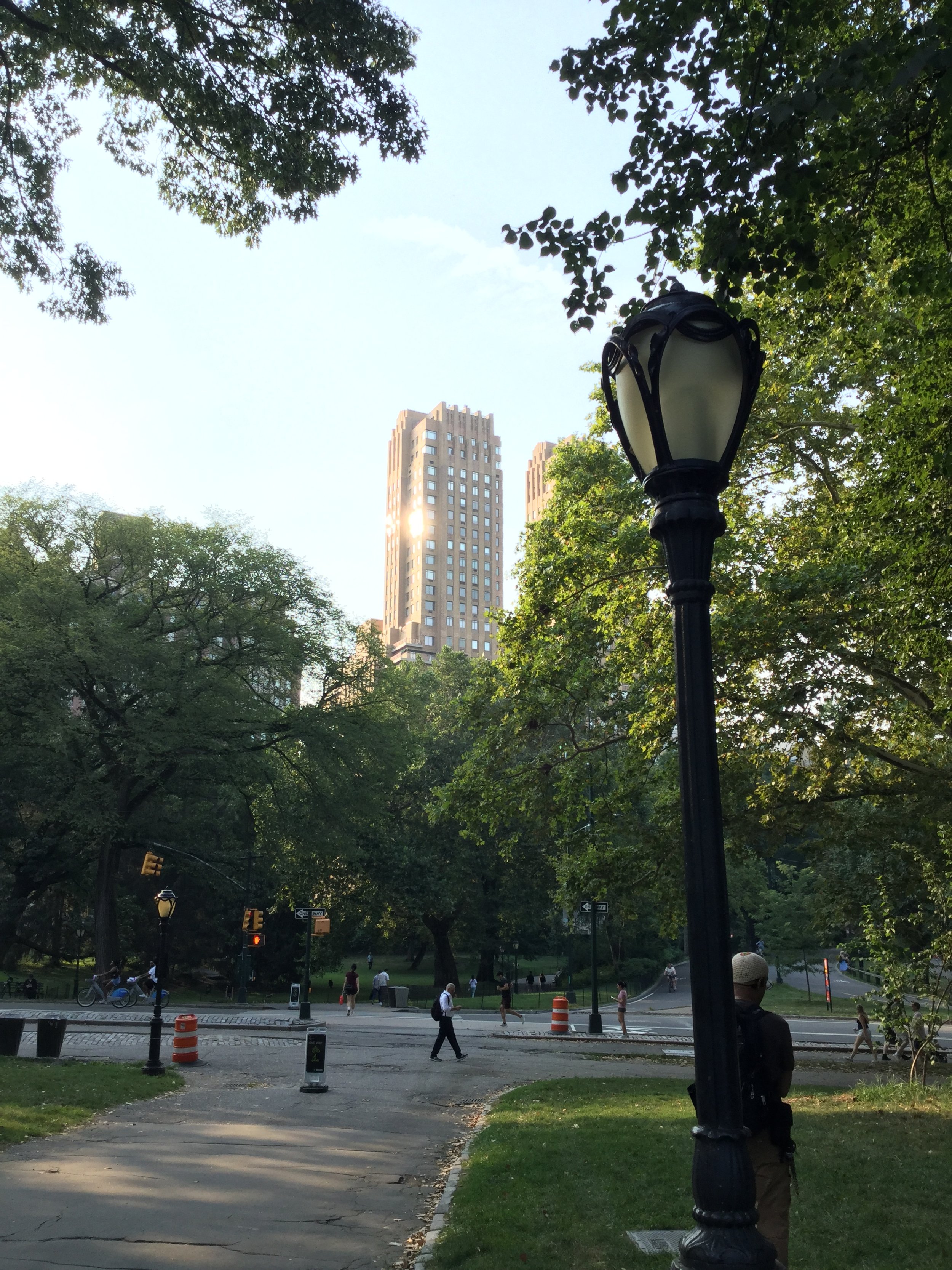 Urban park scene with trees, a street lamp, pedestrians, and tall buildings in the background, taken during daytime.