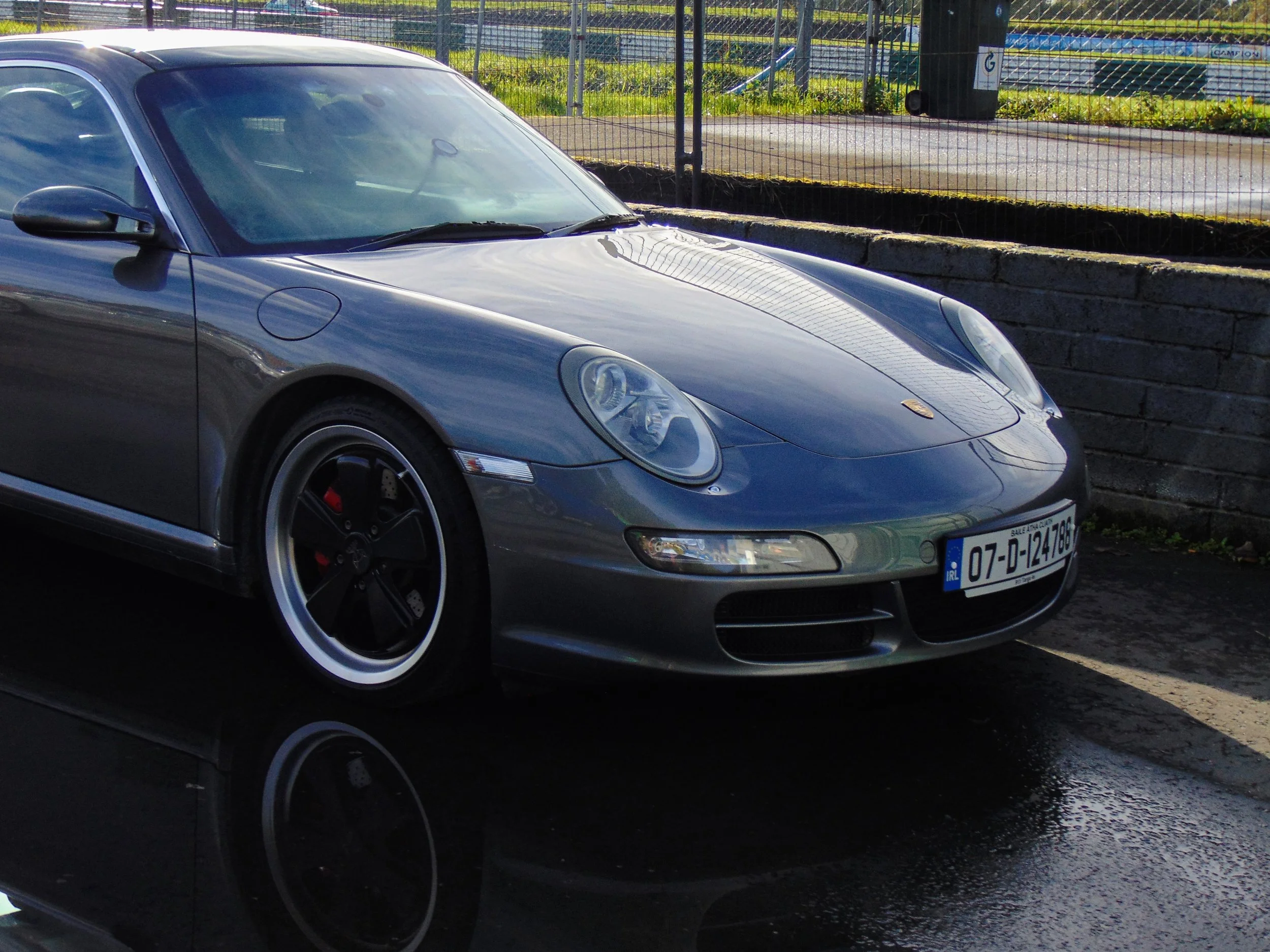 A black Porsche sports car parked on a wet surface near a stone wall and a fence.