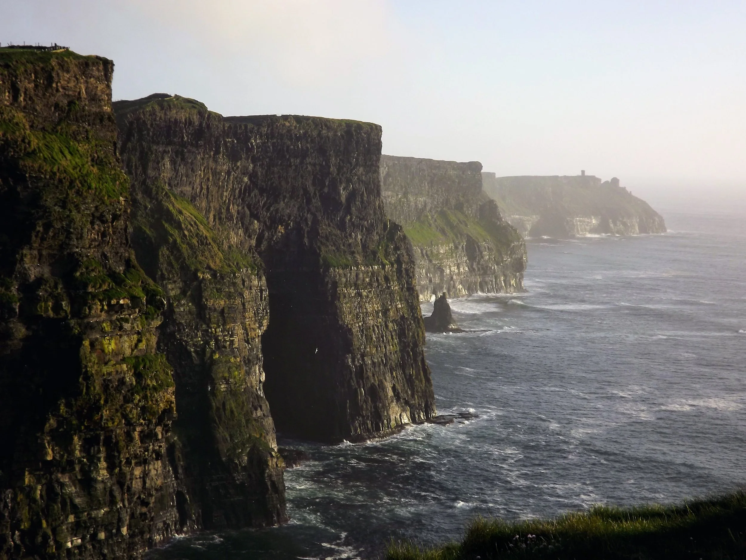 Cliffs along a rugged coastline with the ocean waves crashing at the base, greenery on top of the cliffs, and a distant view of the coastline fading into fog or mist.