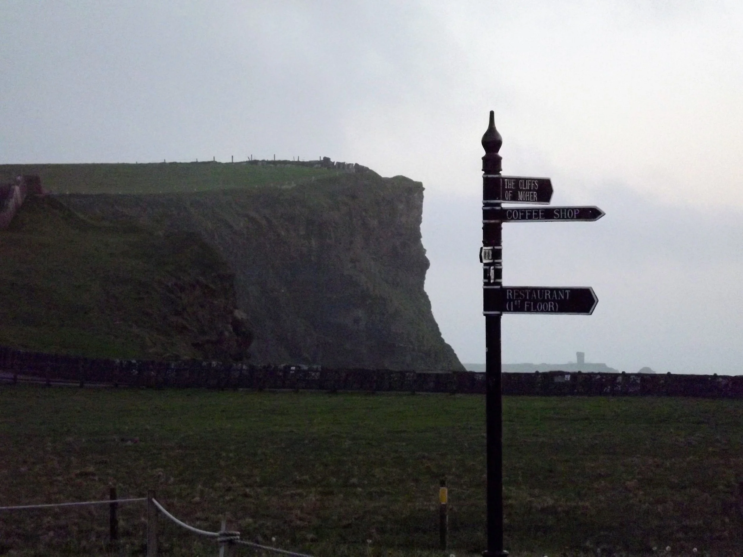 A signpost with multiple directional signs near a grassy area with a cliff in the background. The signs point to the Cliffs of Moher, a coffee shop, and a restaurant on the first floor. The sky appears overcast or foggy.