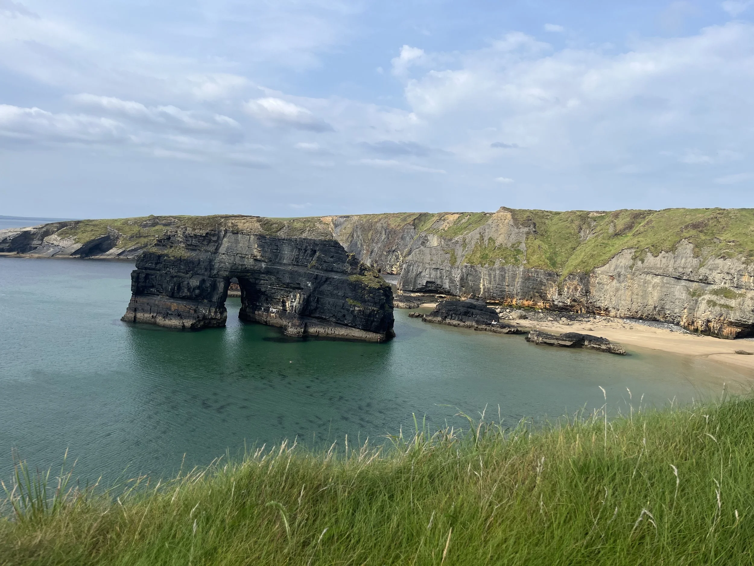 Coastal landscape with green grass in the foreground, large rock formation with an arch in the water, and cliffs with patches of grass under a partly cloudy sky.