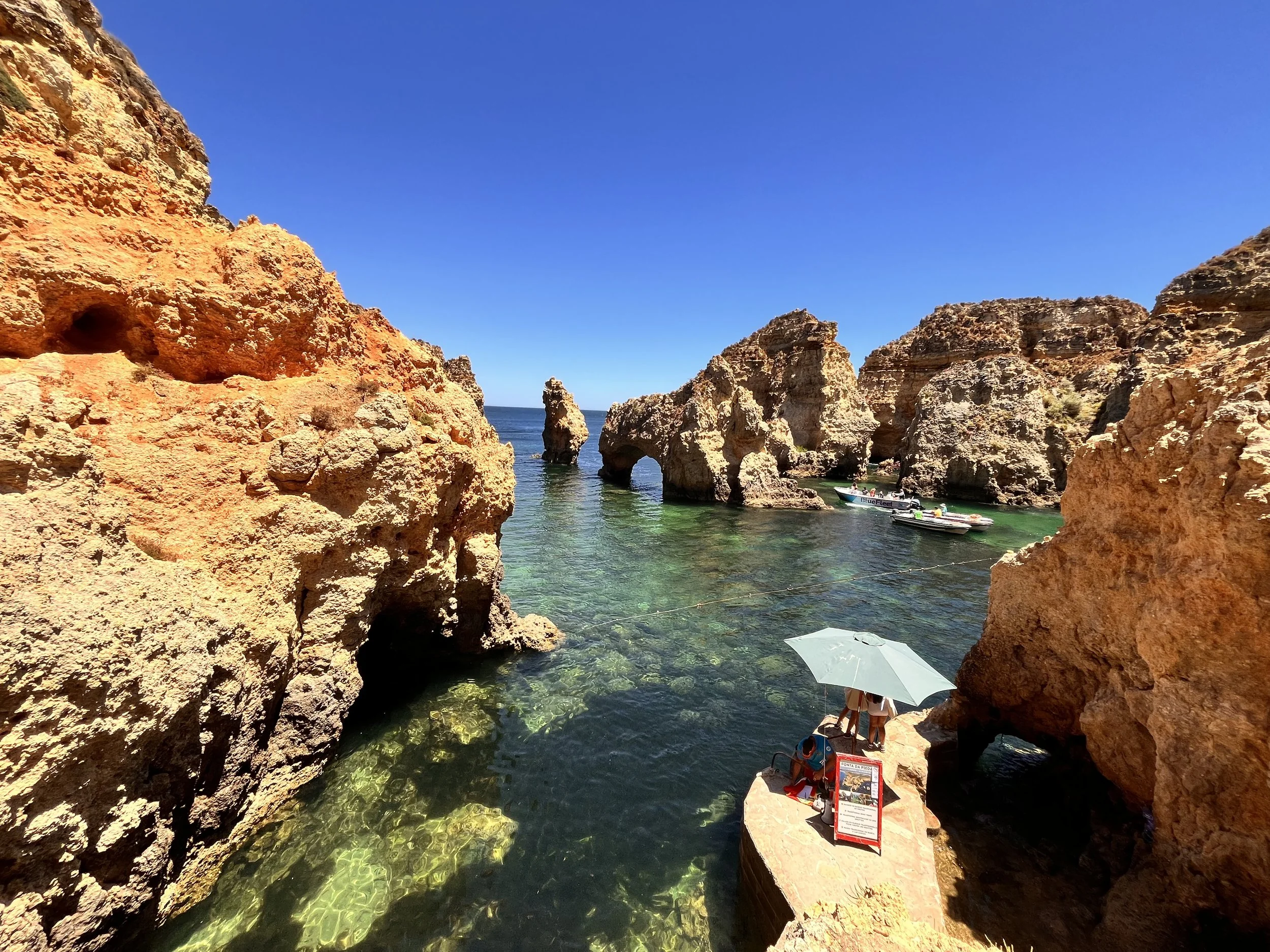 Cliffs overlooking a crystal-clear ocean with boats and a person with an umbrella on a rocky ledge.