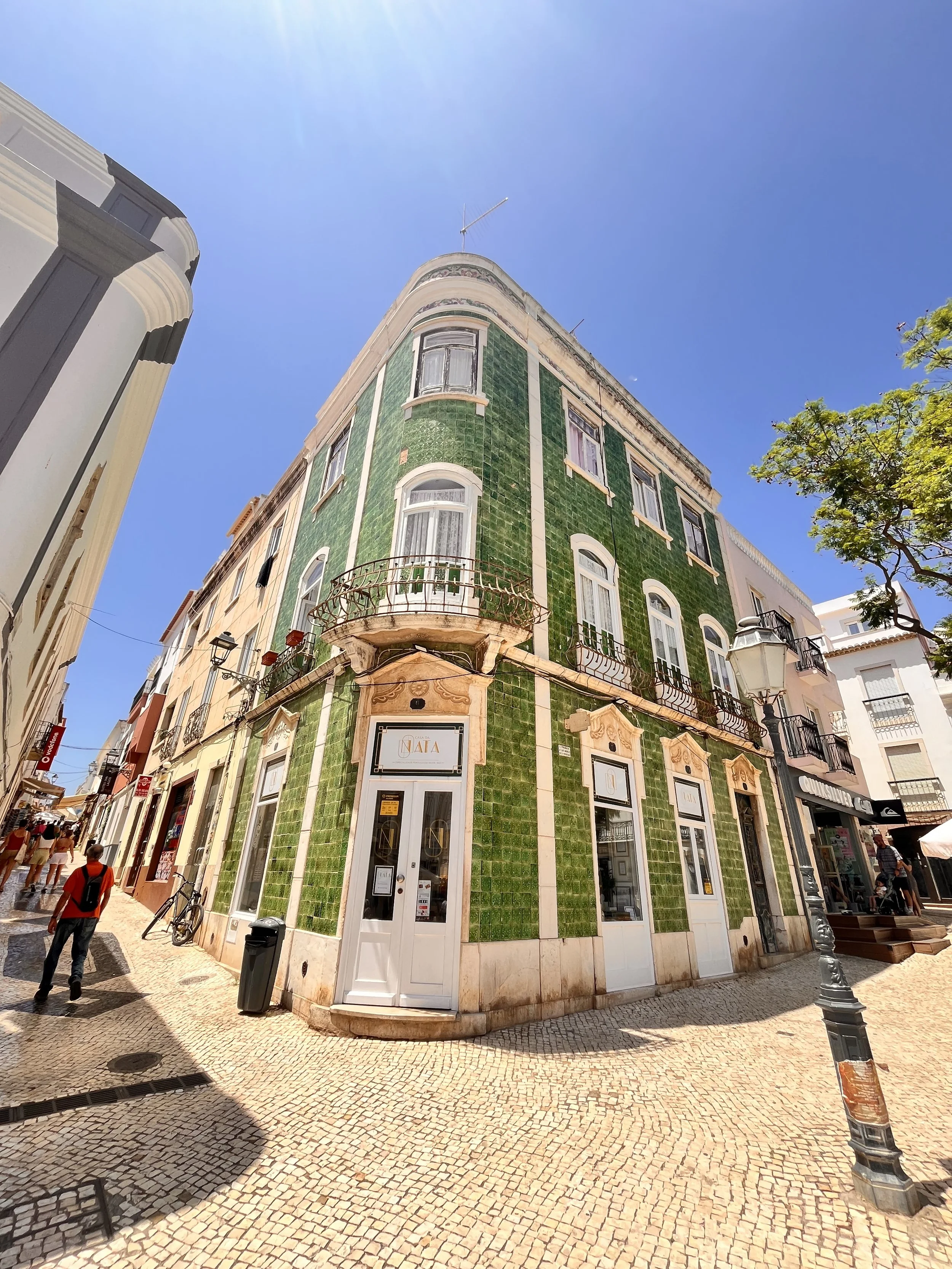 A corner building with green tiled facade on a sunny street with pedestrians, streetlamp, and trees.