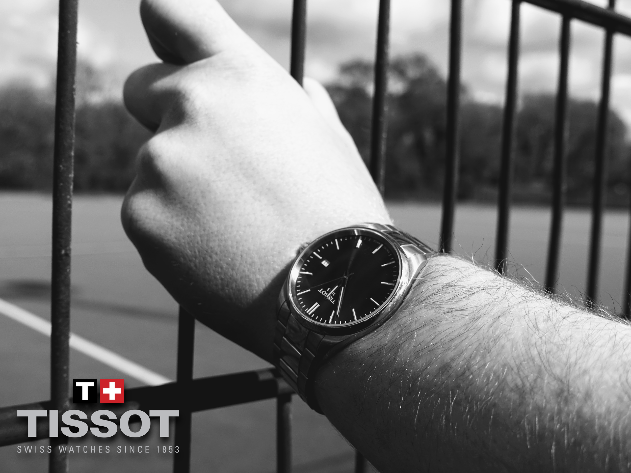 Close-up of a person's arm wearing a wristwatch, reaching through the bars of a fence, with a blurred background.