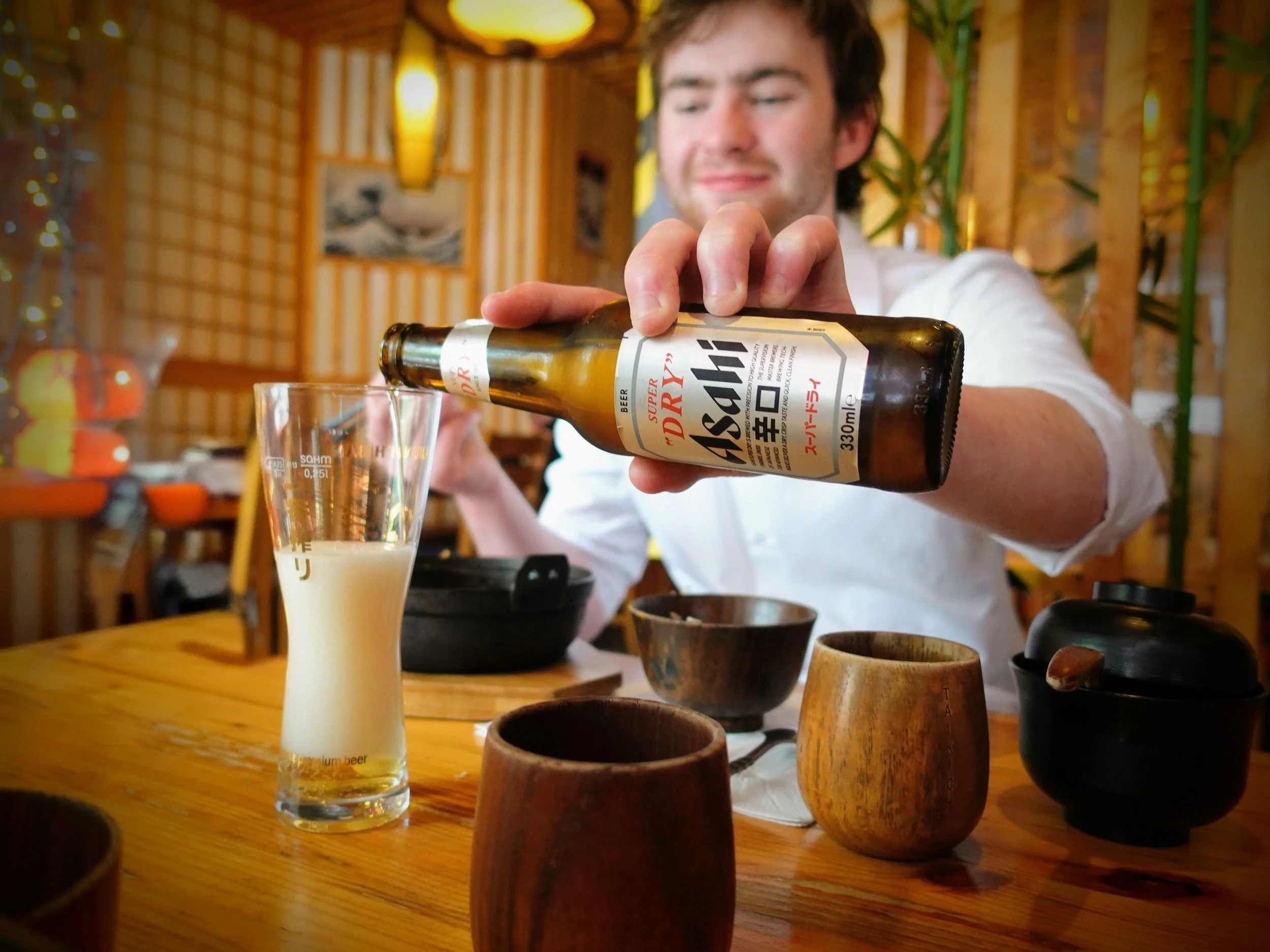 A smiling man is pouring beer from a brown bottle into a tall glass on a wooden table. There are wooden cups and dishes nearby, photo taken in Taikichi, Limerick City