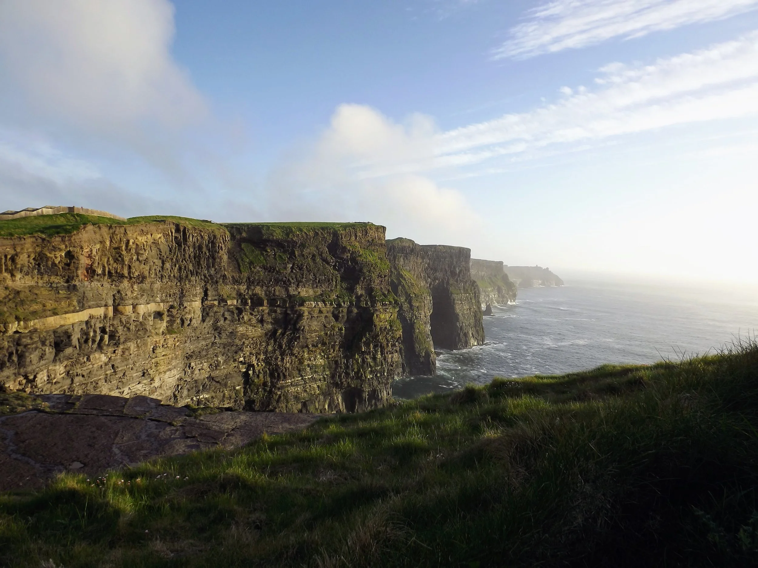 Cliffs along a coastline with grassy top, overlooking the ocean under a partly cloudy sky.