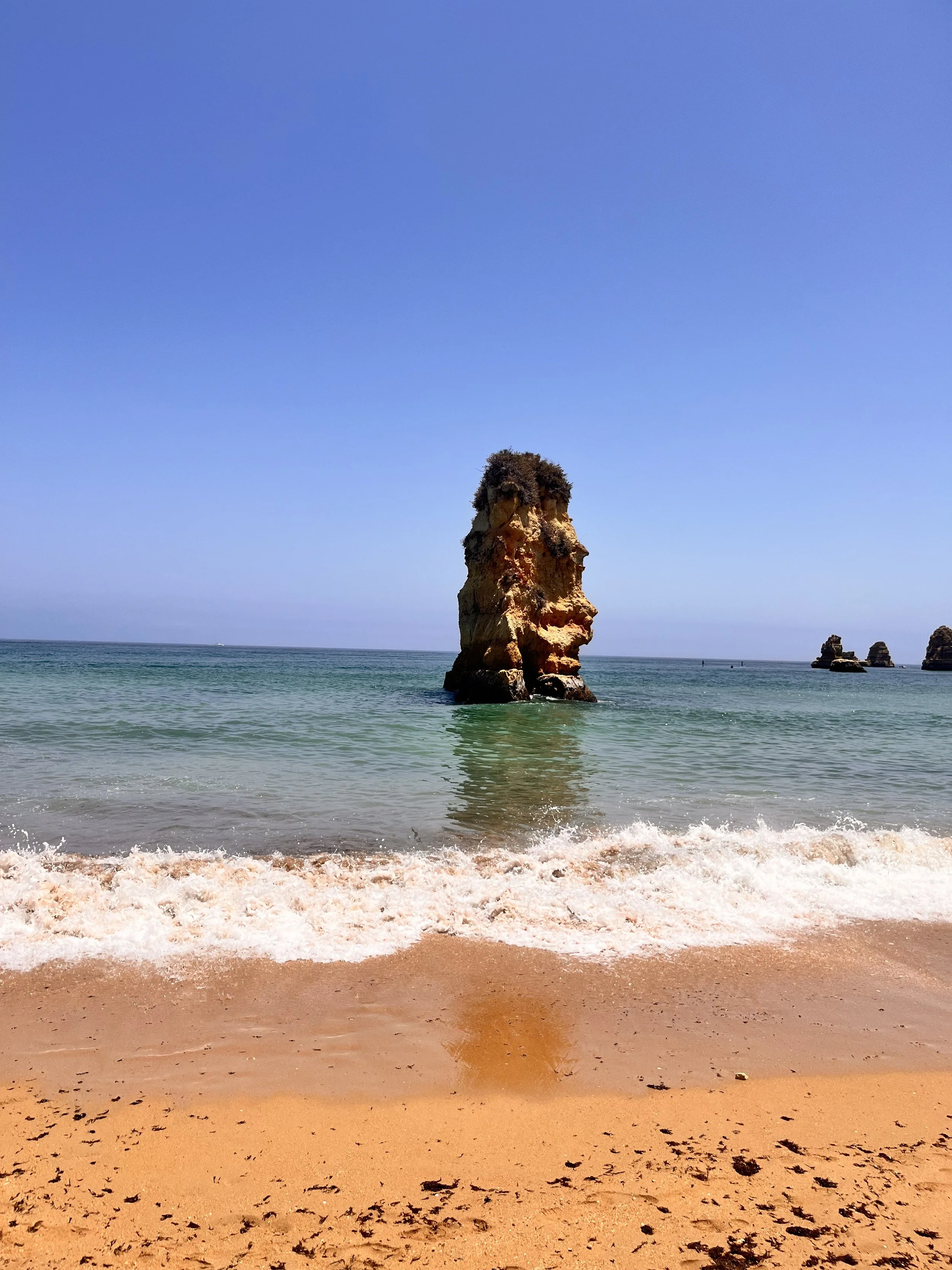 Beach with orange sand, gentle waves, and a rock formation of a large face profile in the water, under a clear blue sky.