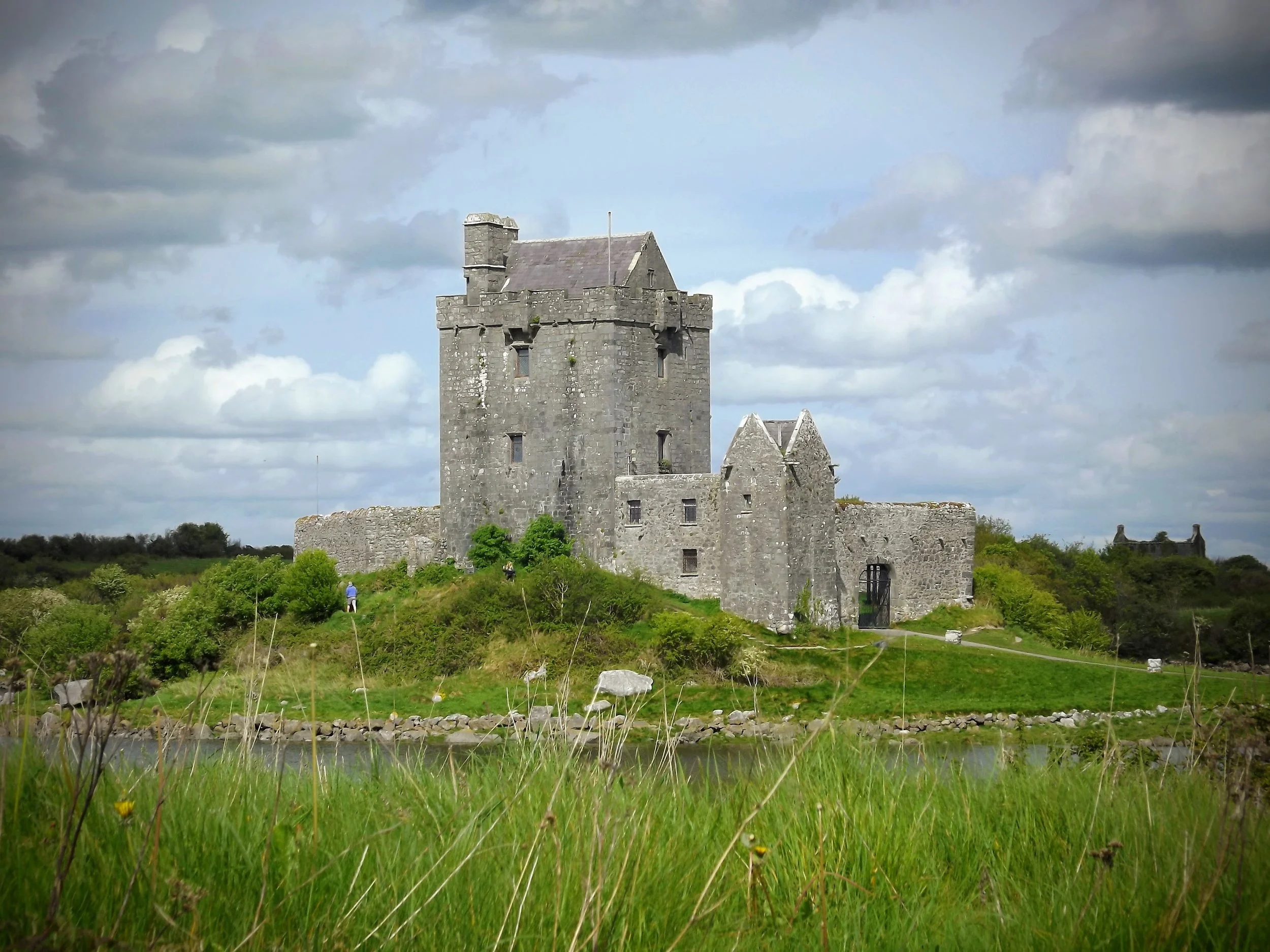 An old stone castle with a tall tower and surrounding walls sits on a grassy hill near a small body of water under a partly cloudy sky.