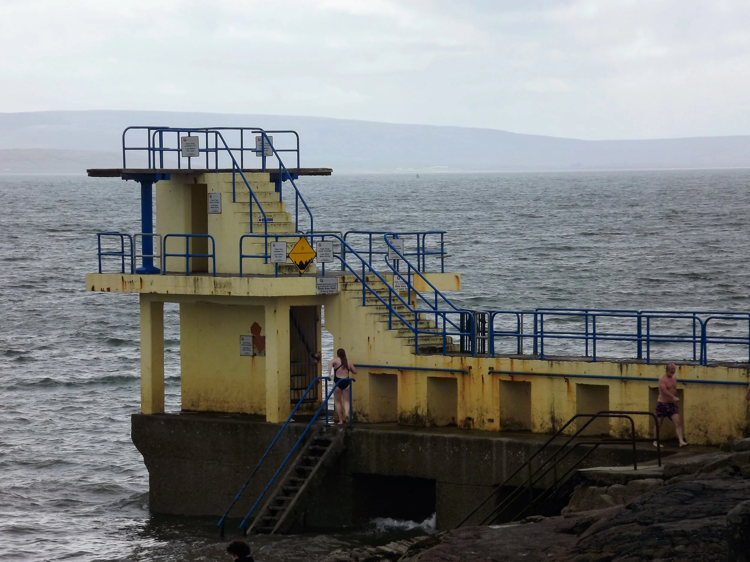 A yellow and blue lifeguard station on a concrete pier extends into a body of water. Two people are near the station, one descending stairs and one walking along the pier. The sky is overcast.