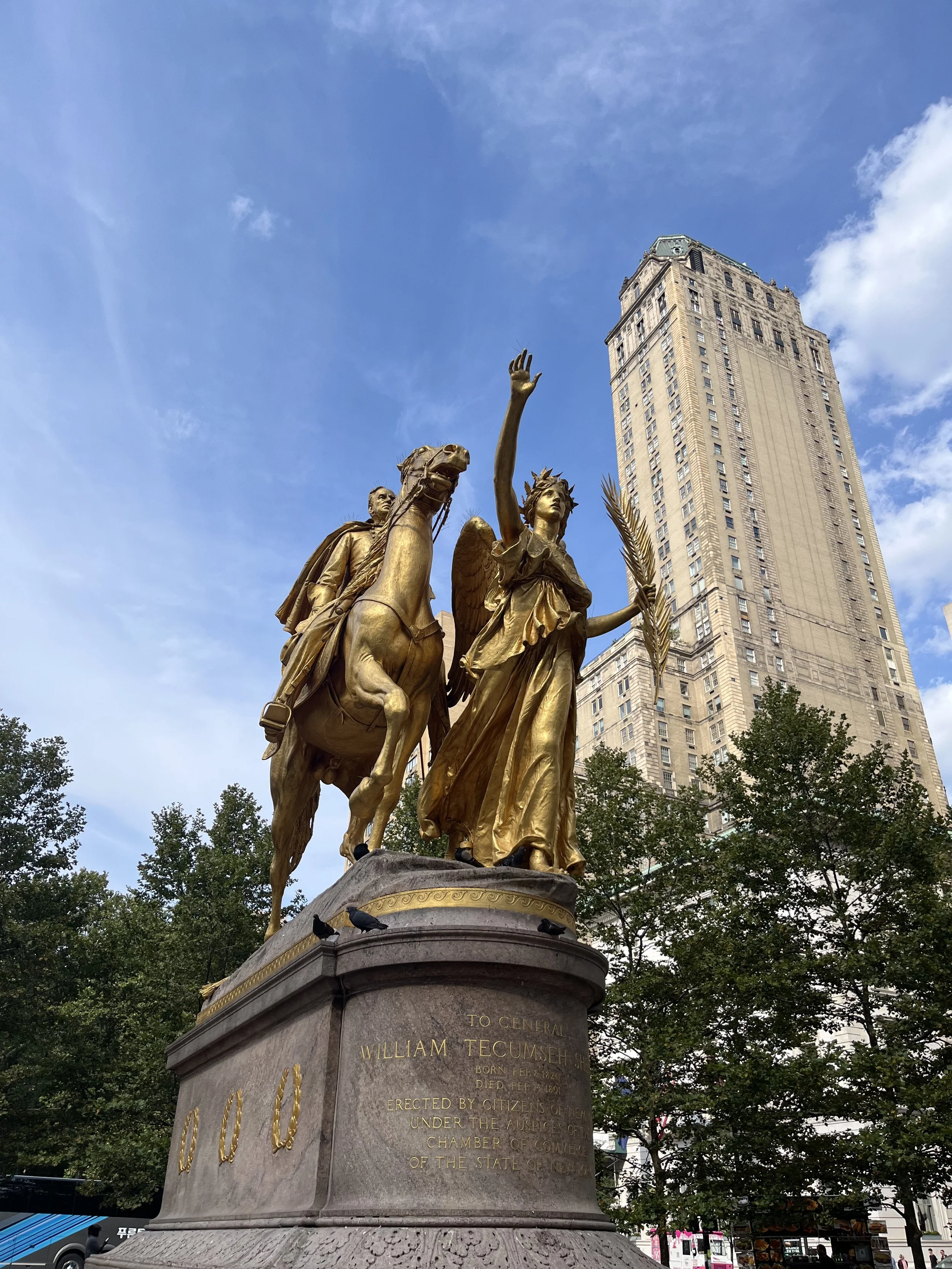 A golden statue of a woman with wings, riding a horse, holding a palm branch, and raising her right hand, with a man riding on the horse behind her. The statue is in a city square with a tall skyscraper and trees in the background.
