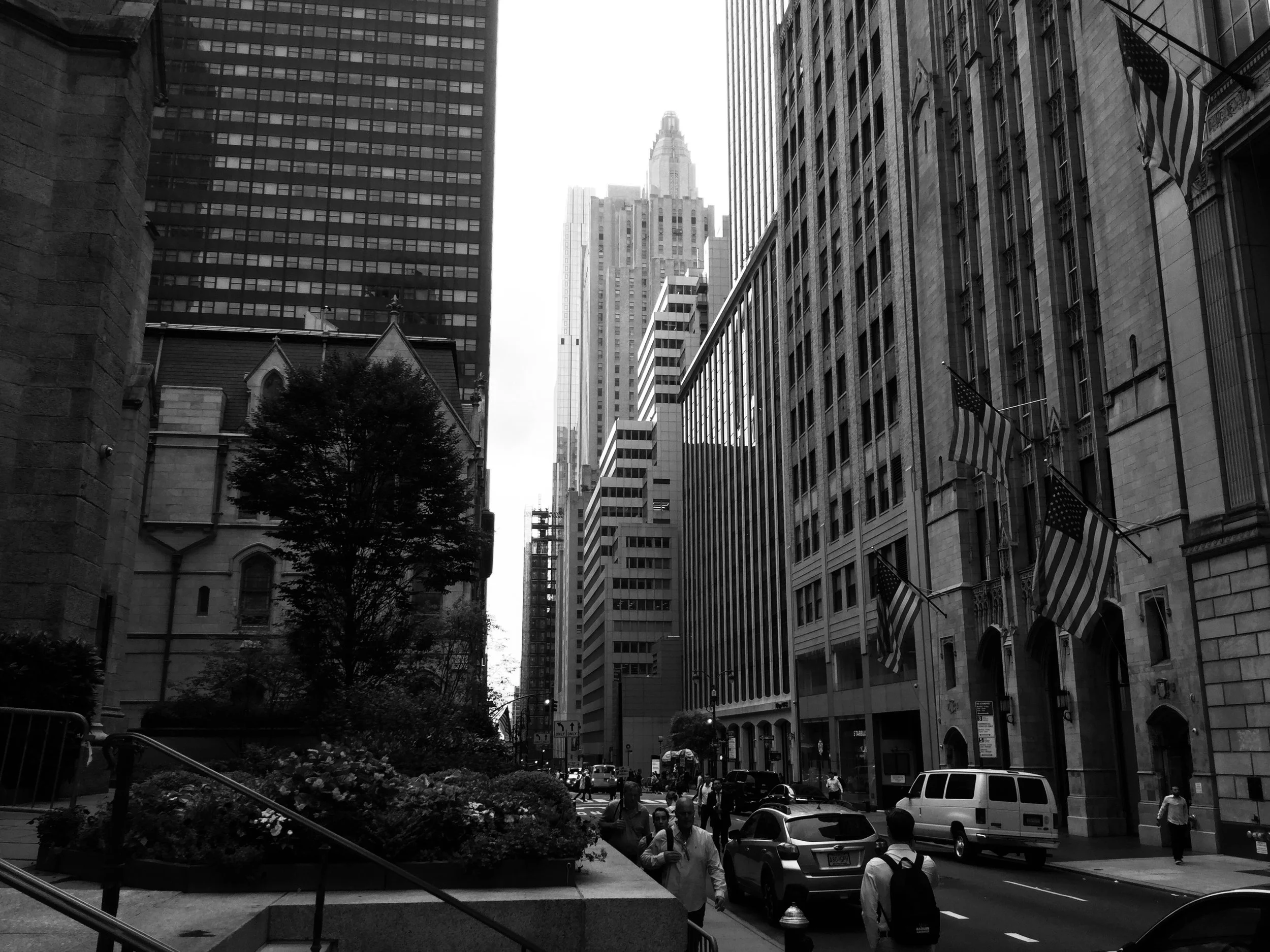 Black and white photo of a city street with tall buildings, flags, and pedestrians.
