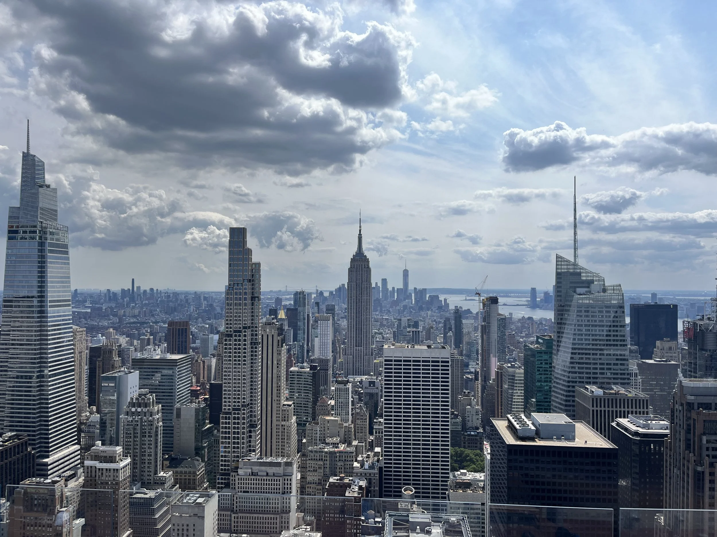 A city skyline featuring tall skyscrapers, including the Empire State Building, with a river in the background and partly cloudy sky.