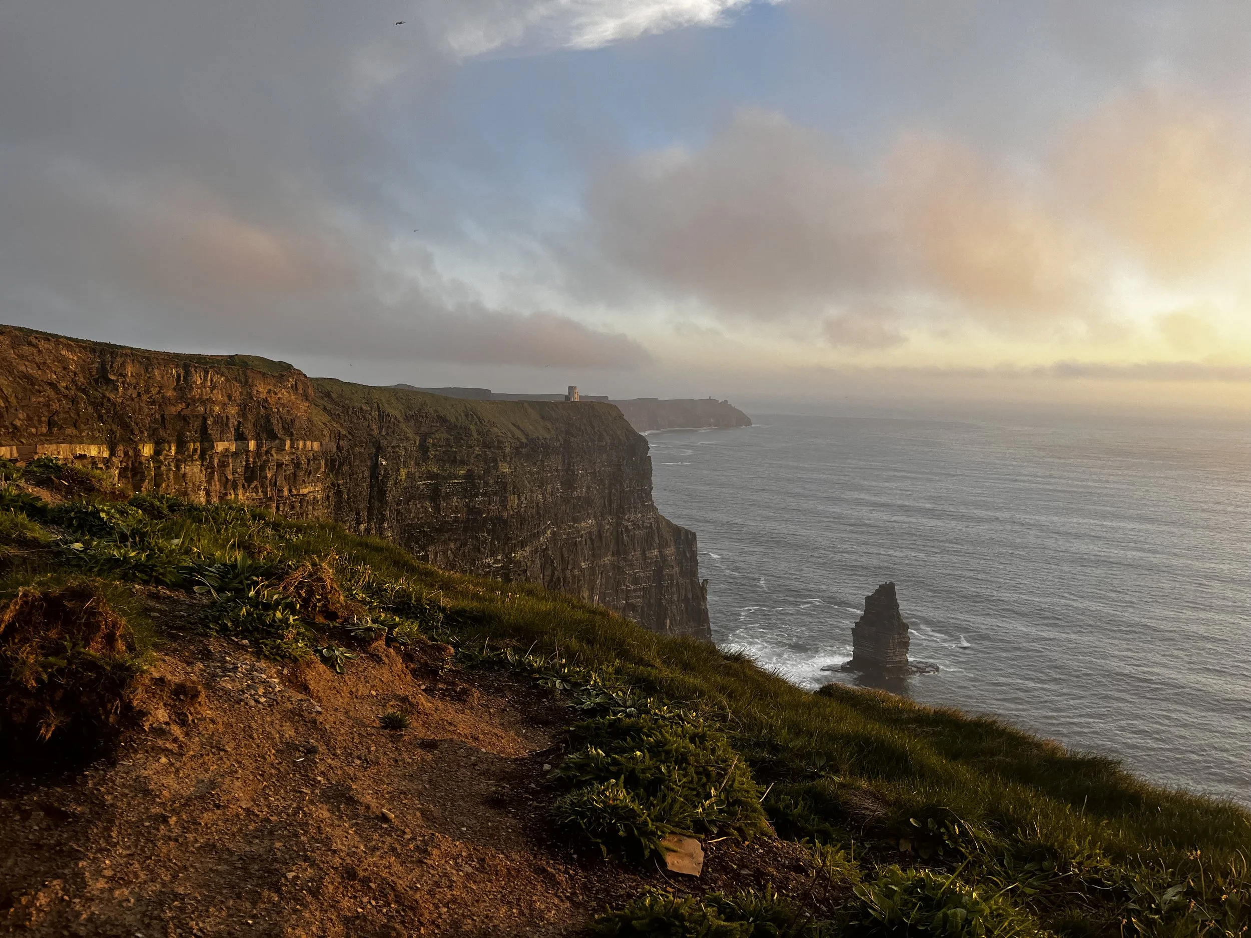 Cliffs along a rugged coastline with a rocky sea stack in the water and a lighthouse far in the distance, under a partly cloudy sky at sunset.