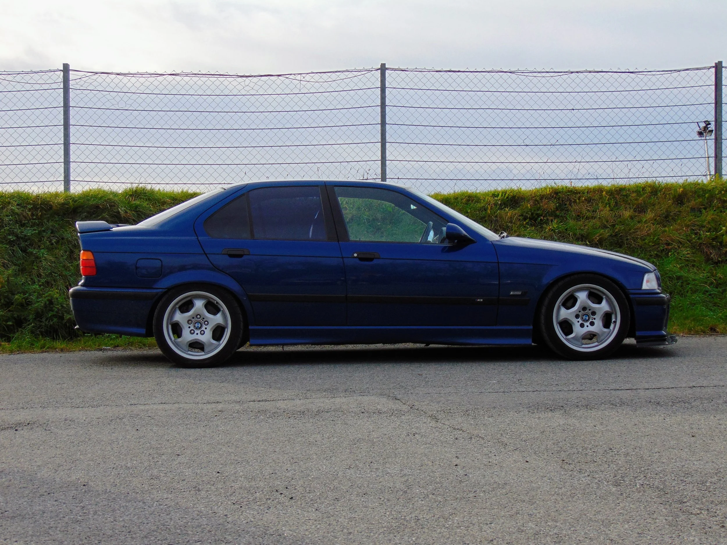 Side view of a blue sedan car parked on a paved road near a grassy embankment with a chain-link fence in the background.
