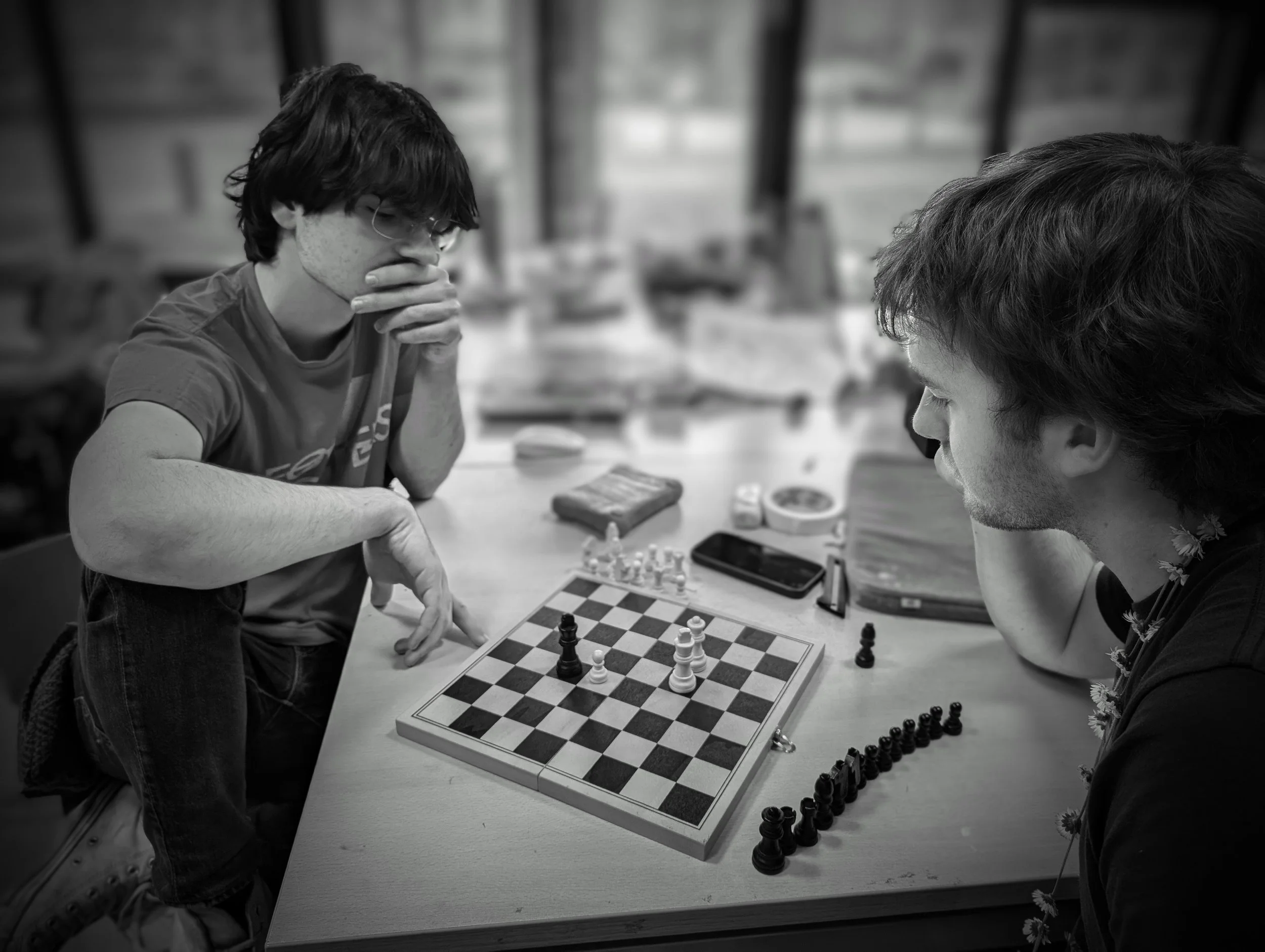Two young men playing chess at a table, one is wearing glasses and appears deep in thought while the other looks at the chessboard, both focused on the game.