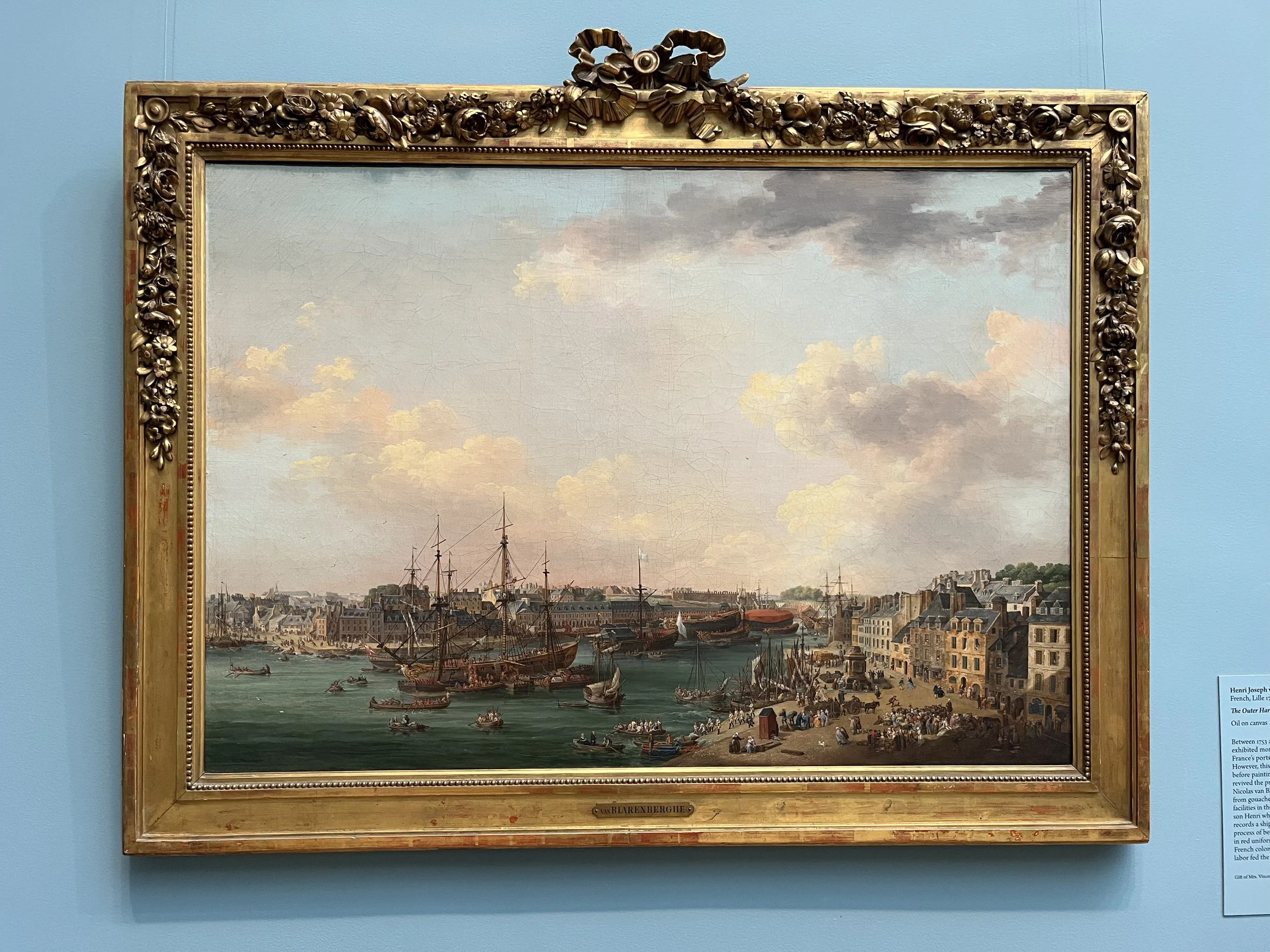 A painting of a bustling harbor scene with ships and boats docked along the waterfront, under a partly cloudy sky. The scene includes historic buildings lining the harbor and many people engaged in various activities.