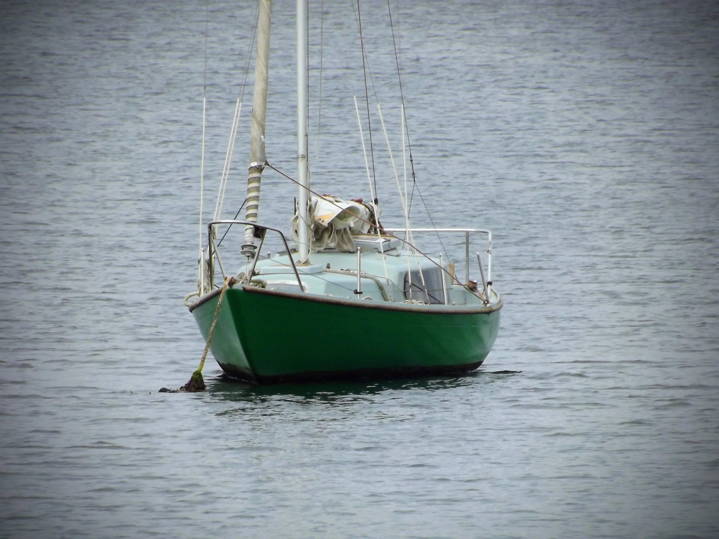 A green sailboat floating on calm water with no visible crew onboard.