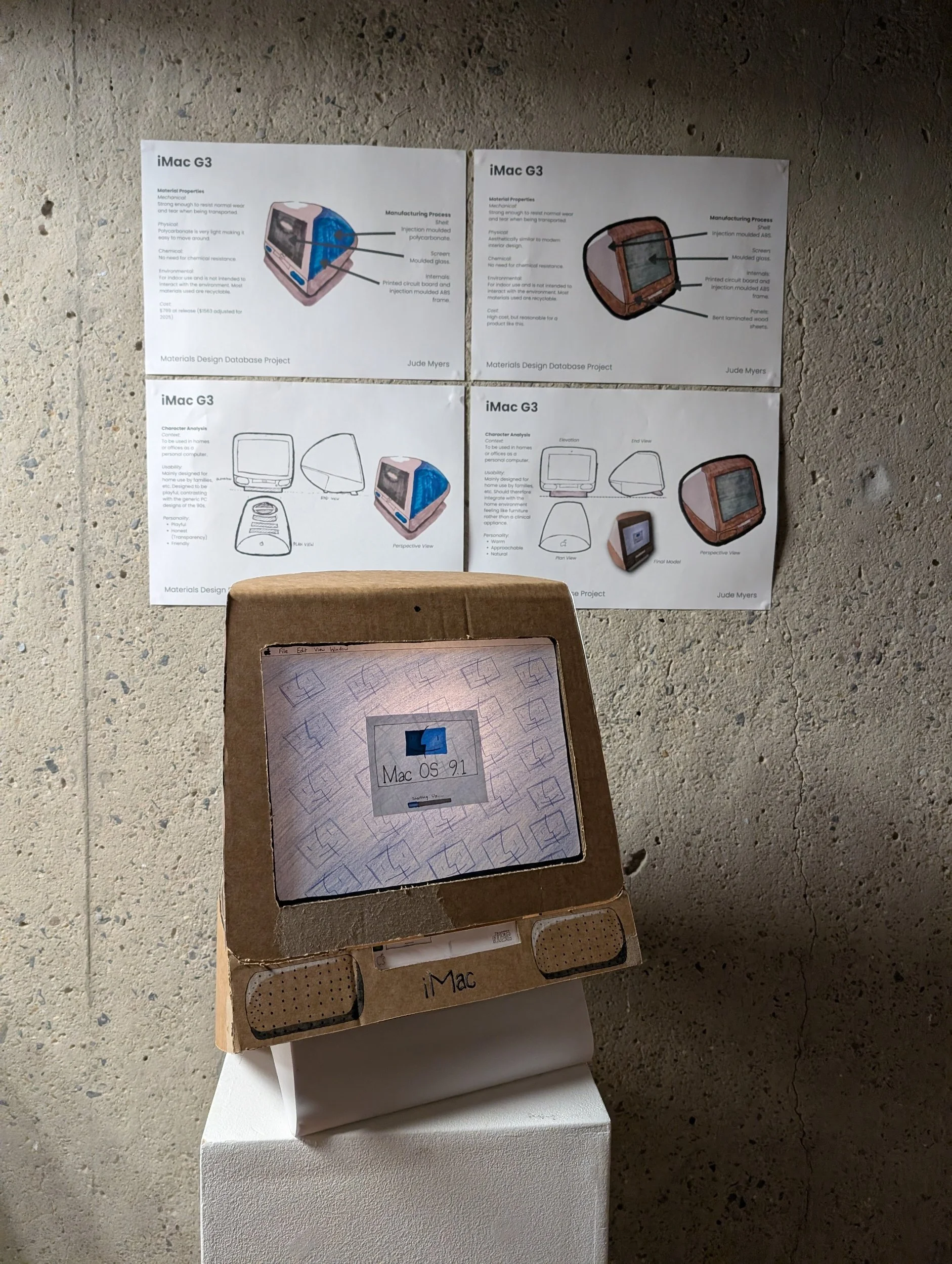 A cardboard display case with a small screen showing the Mac OS 9.1 logo, placed on a white pedestal. Behind it, documents with diagrams and descriptions of an iMac G3 are taped to a concrete wall.