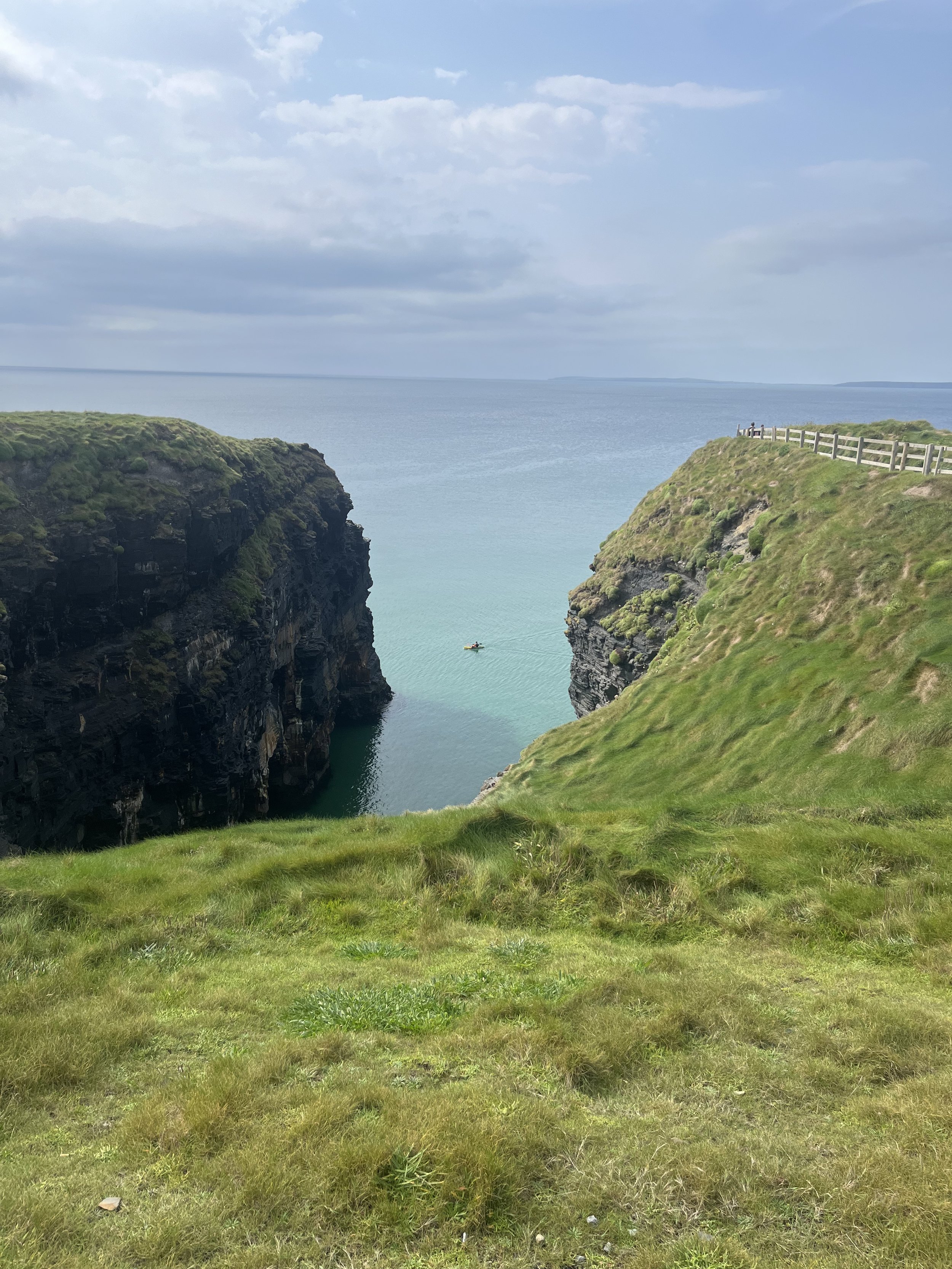 View of a narrow ocean inlet between two grassy cliffs with a small boat sailing in the water, a wooden fence on the right cliff, and a cloudy sky.
