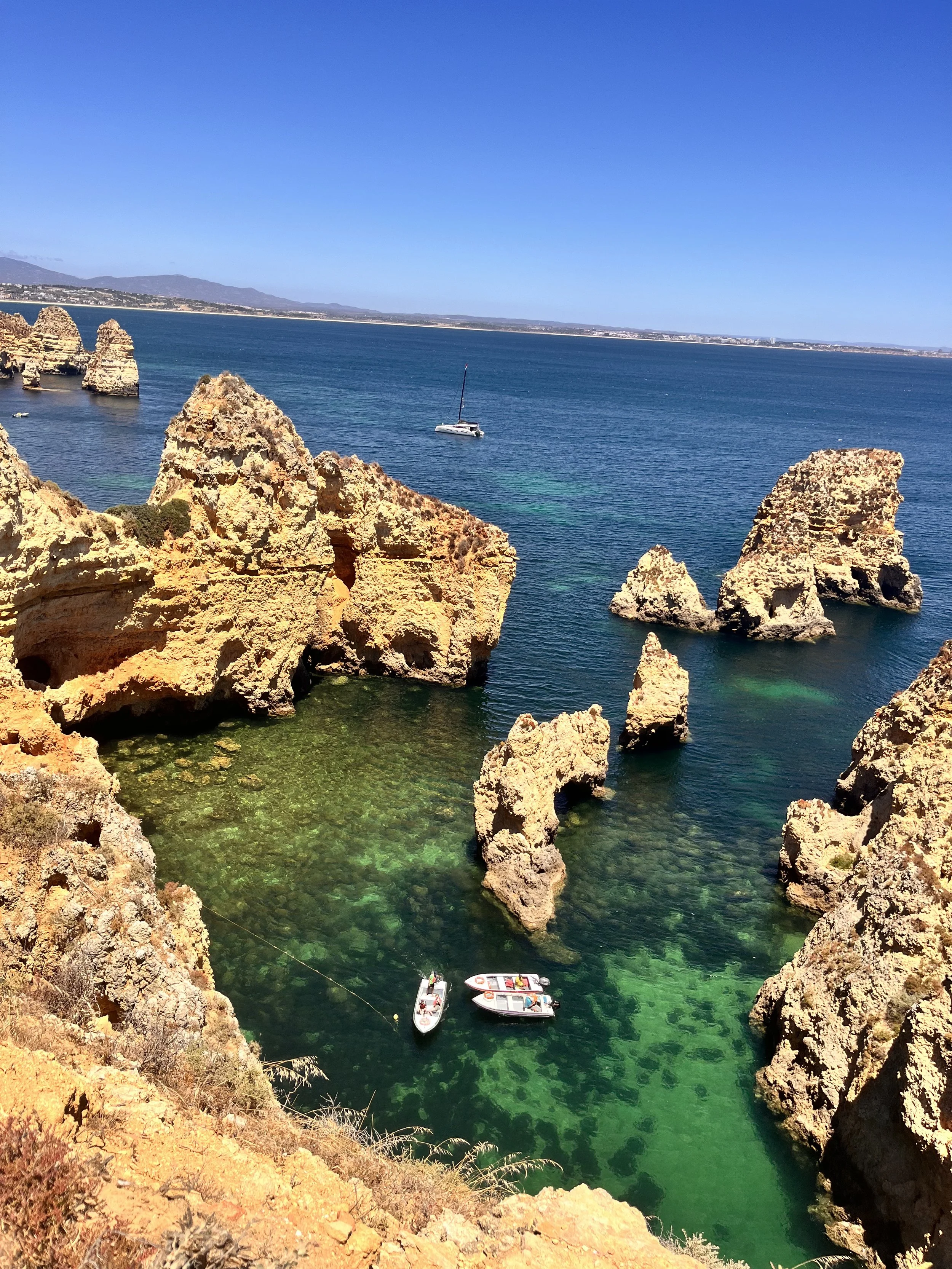 Coastal scene featuring large rocky formations near the shore with boats anchored in clear, green water and a distant horizon with mountains under a bright blue sky.