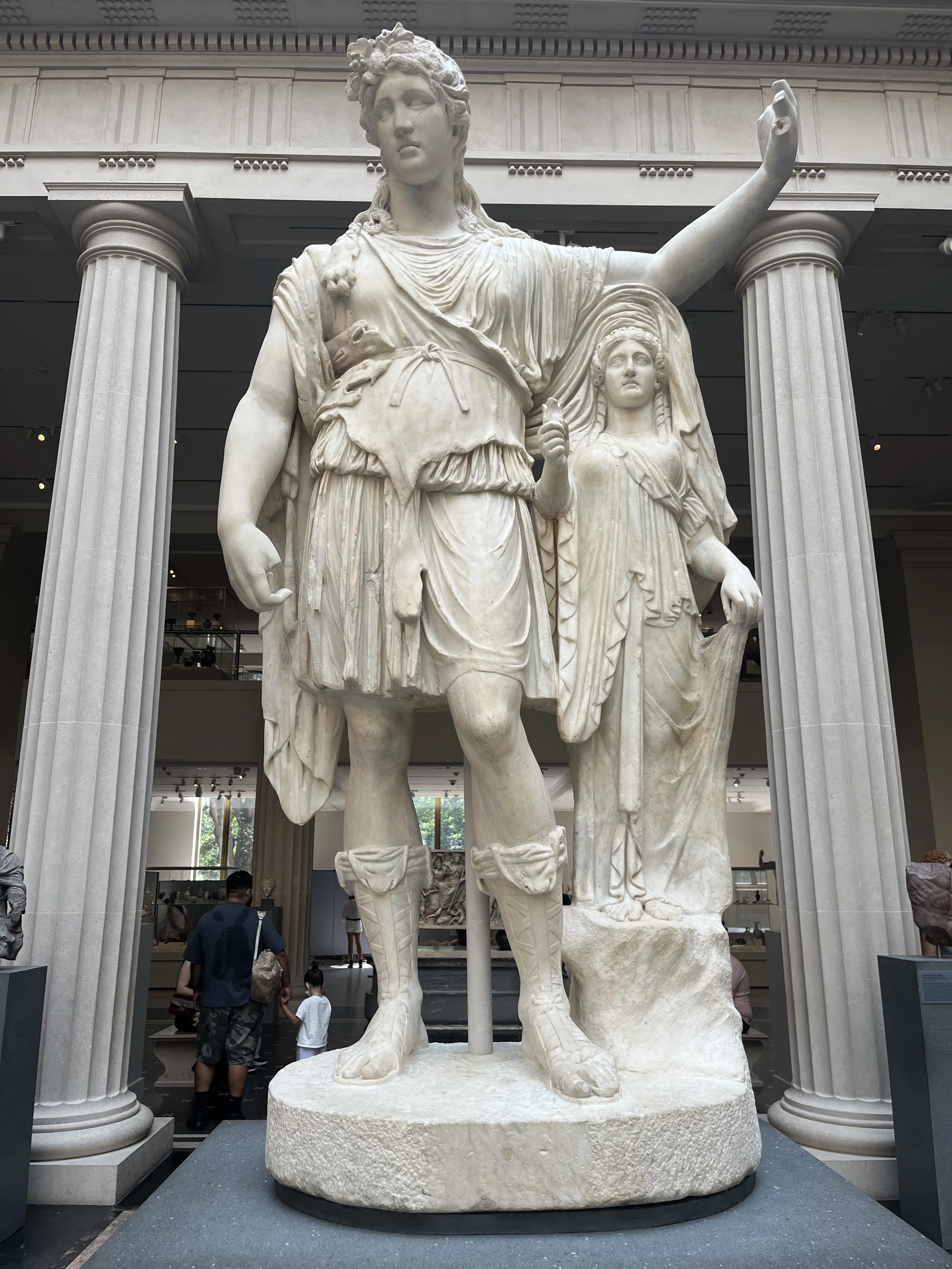 Ancient Greek marble statue of a hero and a girl, with classical columns in the background, displayed in a museum.
