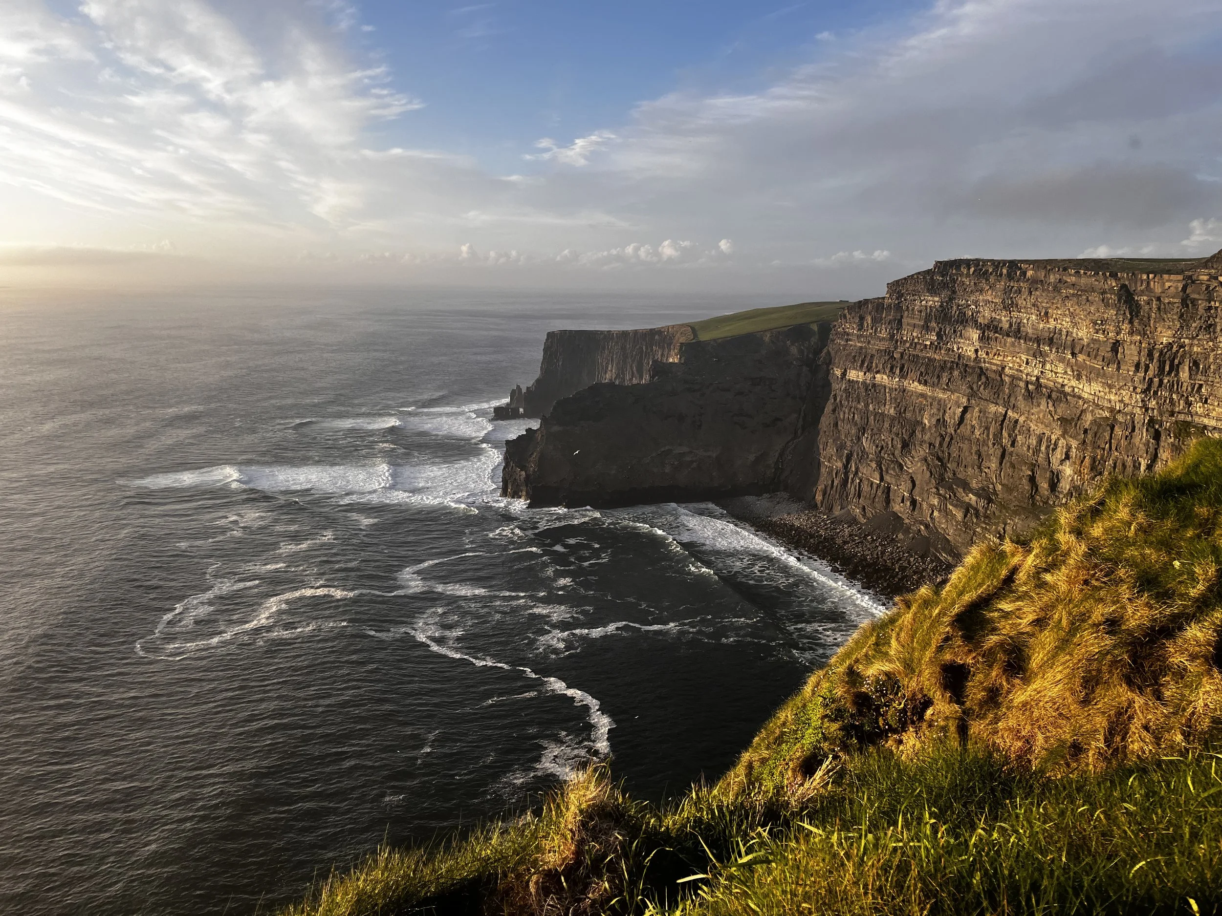 Cliffs overlooking the ocean during sunset with grassy foreground and partly cloudy sky.
