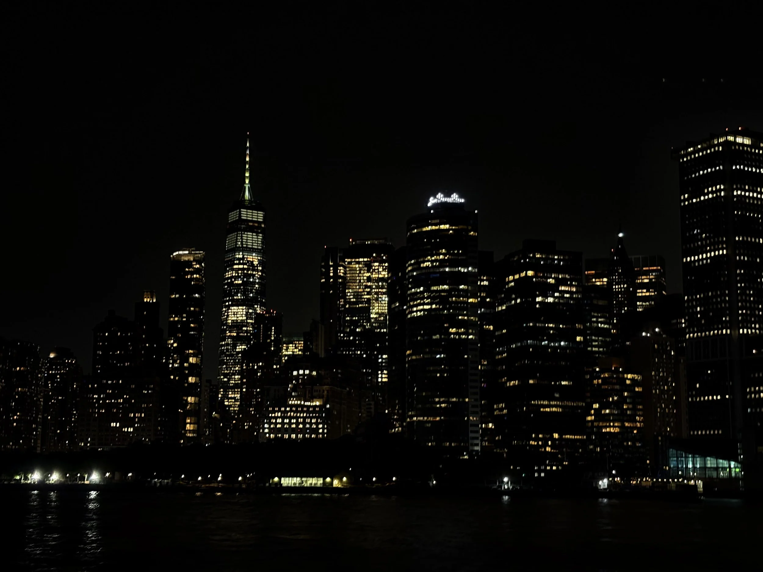 Nighttime view of a city skyline with illuminated skyscrapers, including the One World Trade Center, reflecting on the water.