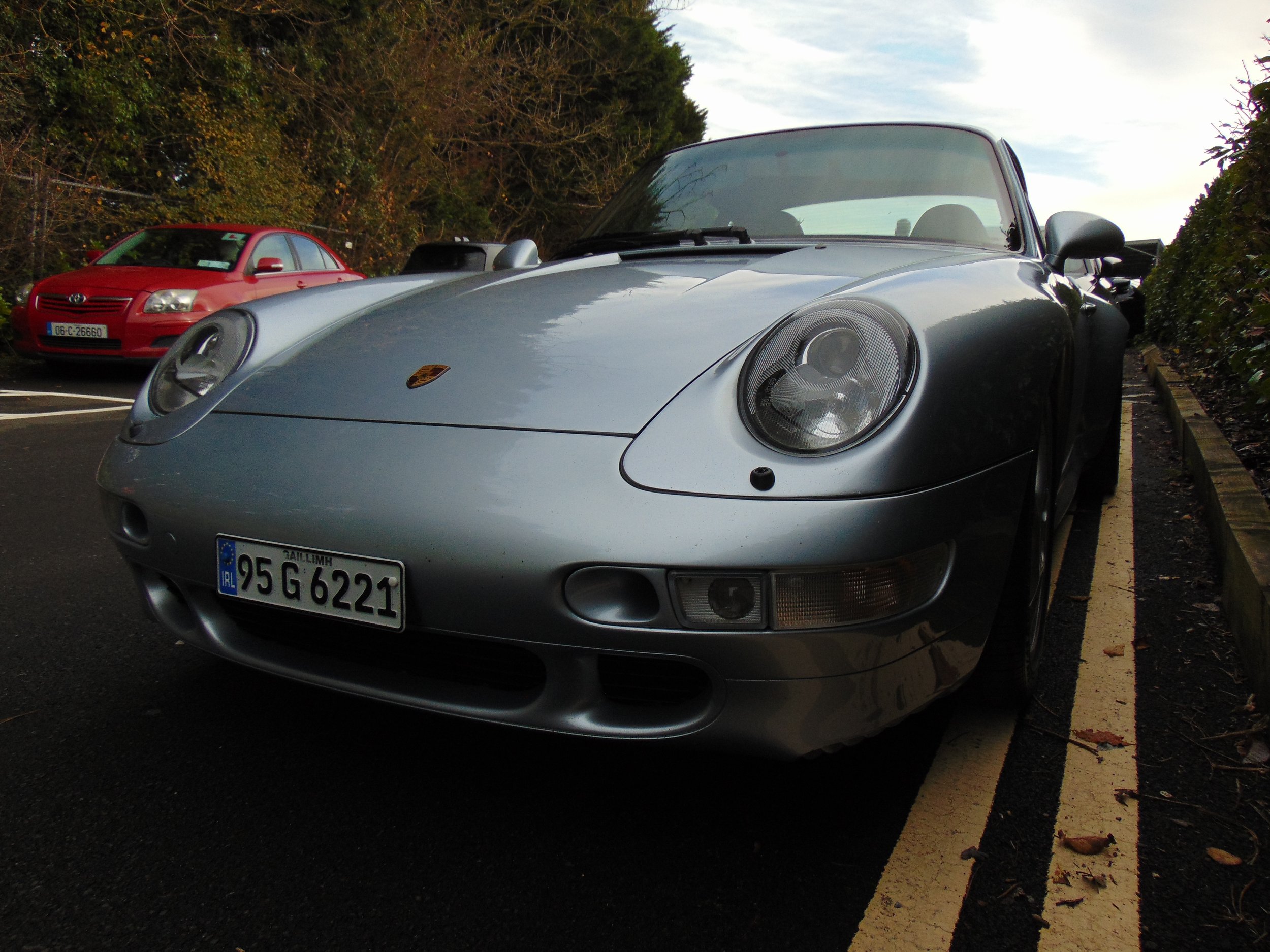 A silver Porsche 911 convertible parked on a parking lot with a red car in the background, trees, and cloudy sky.