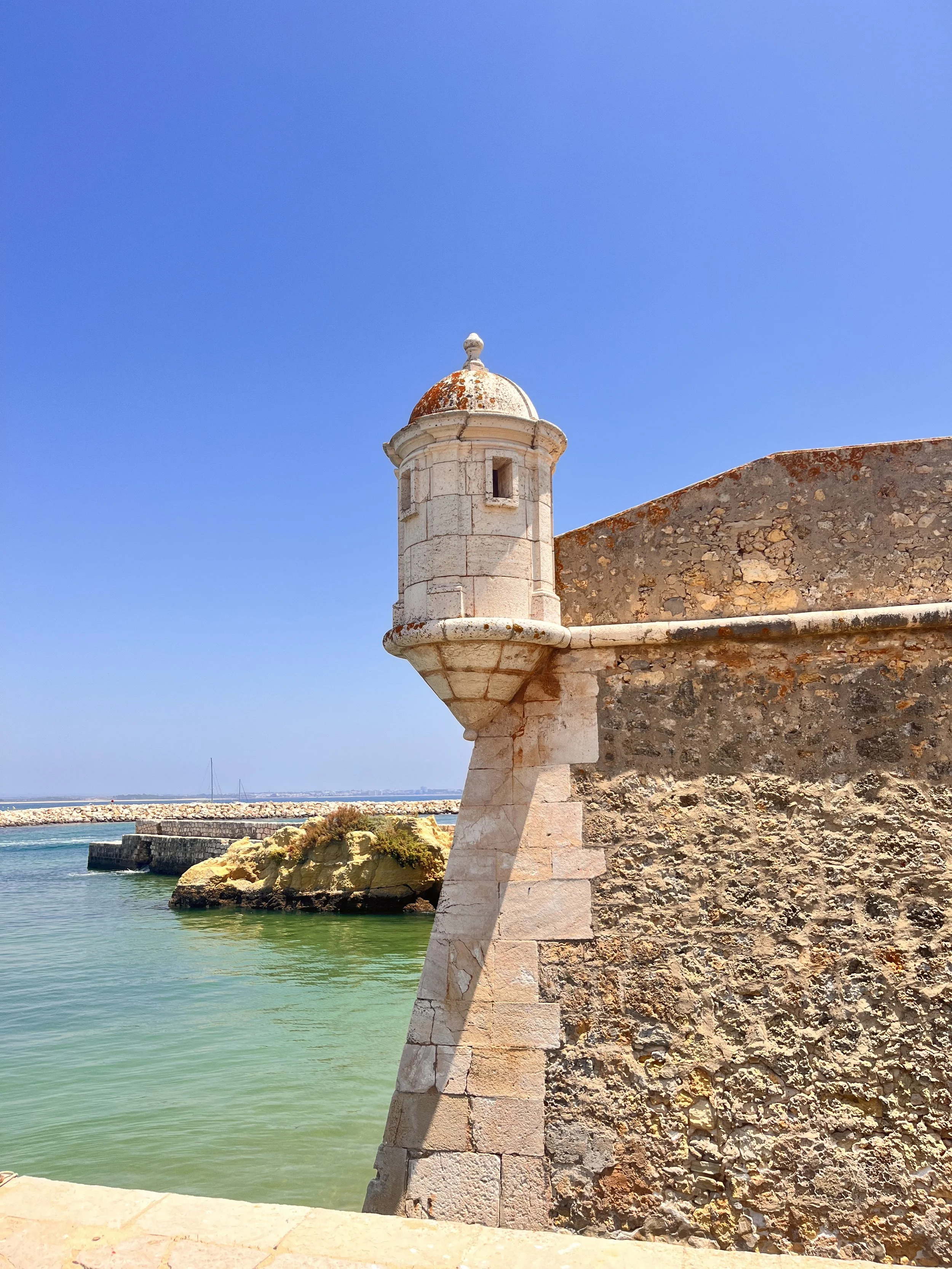 A historic stone fortress with a small round watchtower featuring a domed roof, situated along a waterfront with clear green water, rocky outcroppings, and a bright blue sky.
