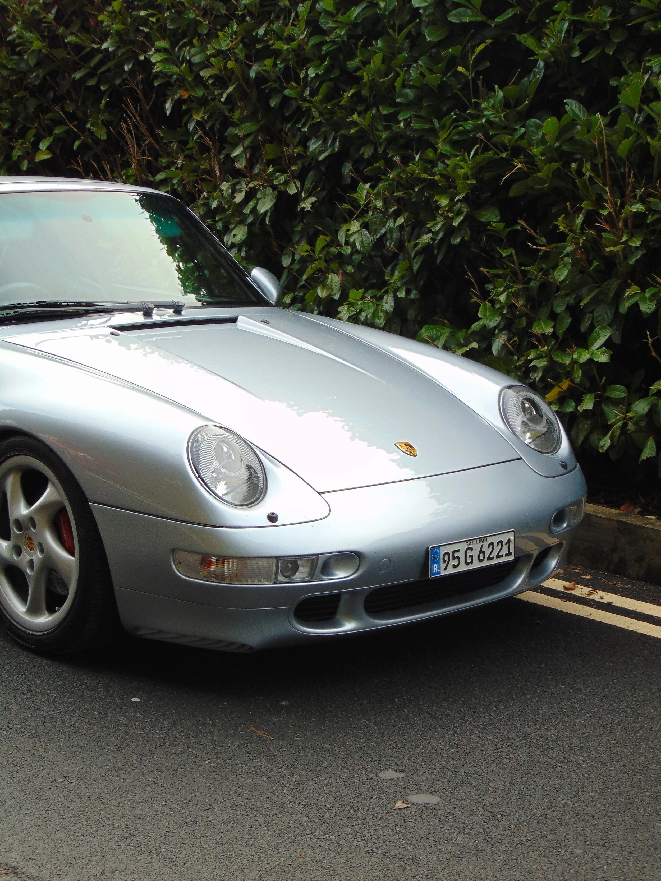 A silver Porsche sports car parked on the street near leafy bushes.