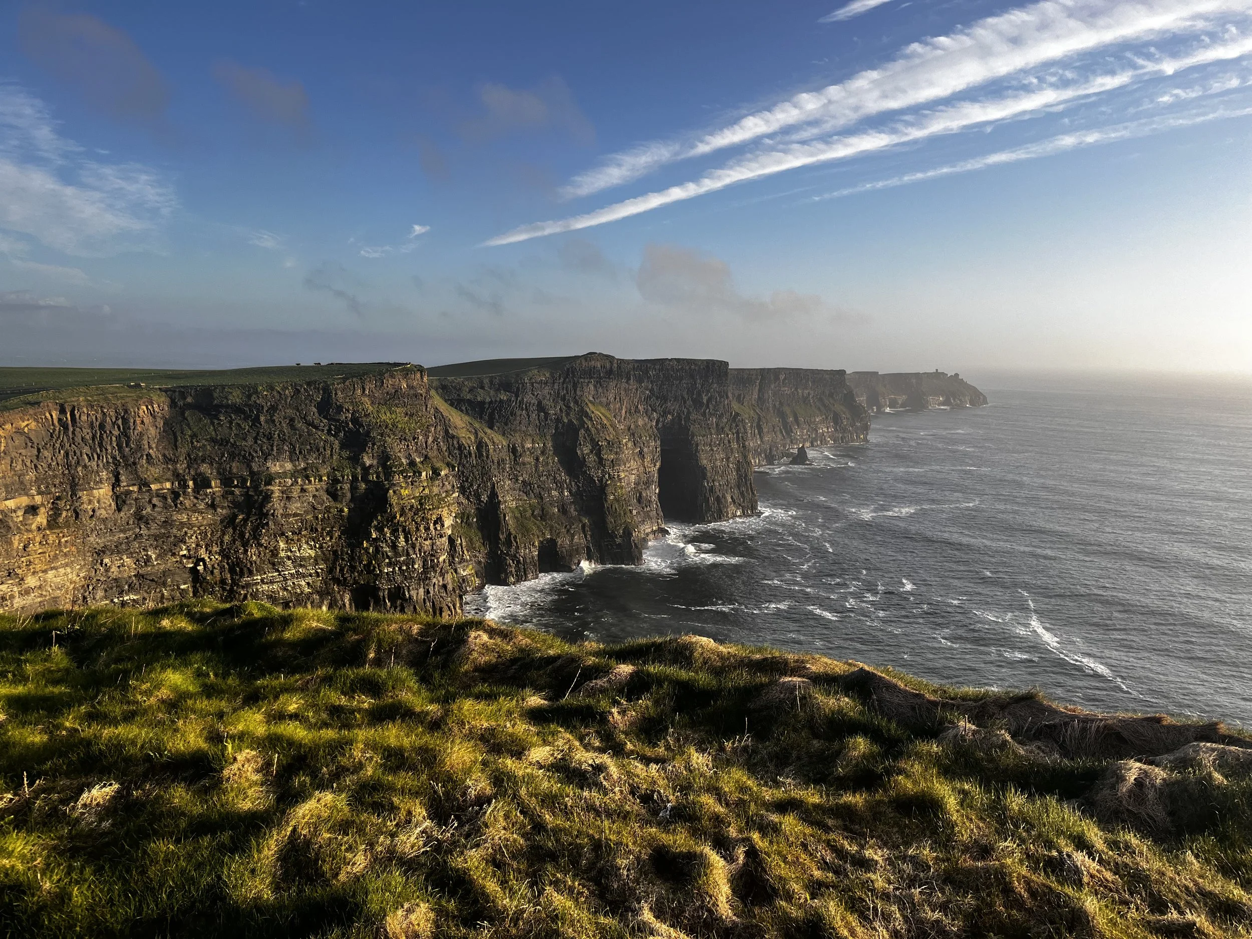 Cliffs overlooking the ocean with grassy foreground and partly cloudy sky.