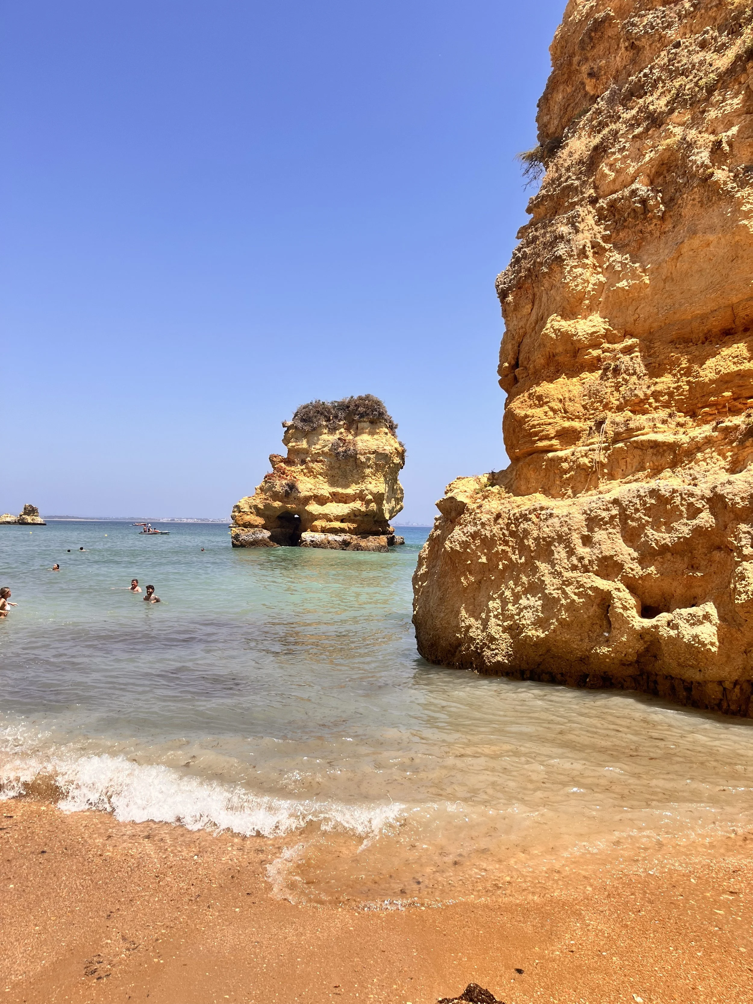 Beach scene with large orange rock formations, clear water, and people swimming.