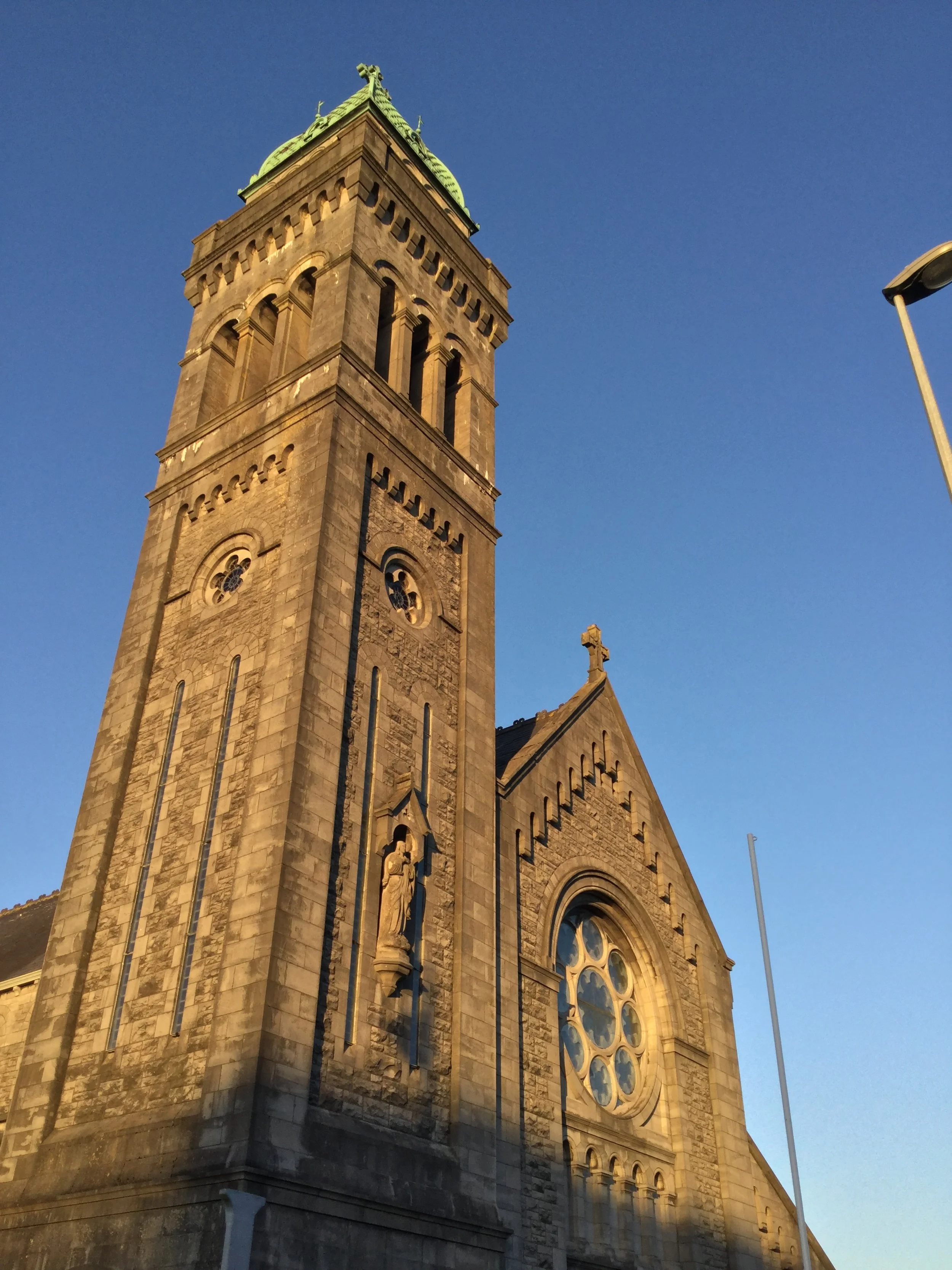 A historic church with a tall clock tower and a large circular stained glass window, bathed in warm sunlight, against a clear blue sky.