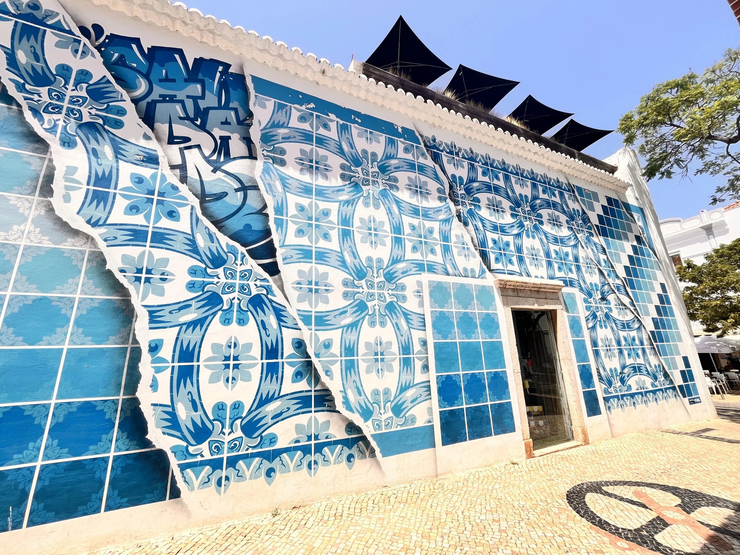 Colorful mural with blue and white tile patterns on a building wall, appearing as a mosaic design, partially torn to reveal the tiles underneath.
