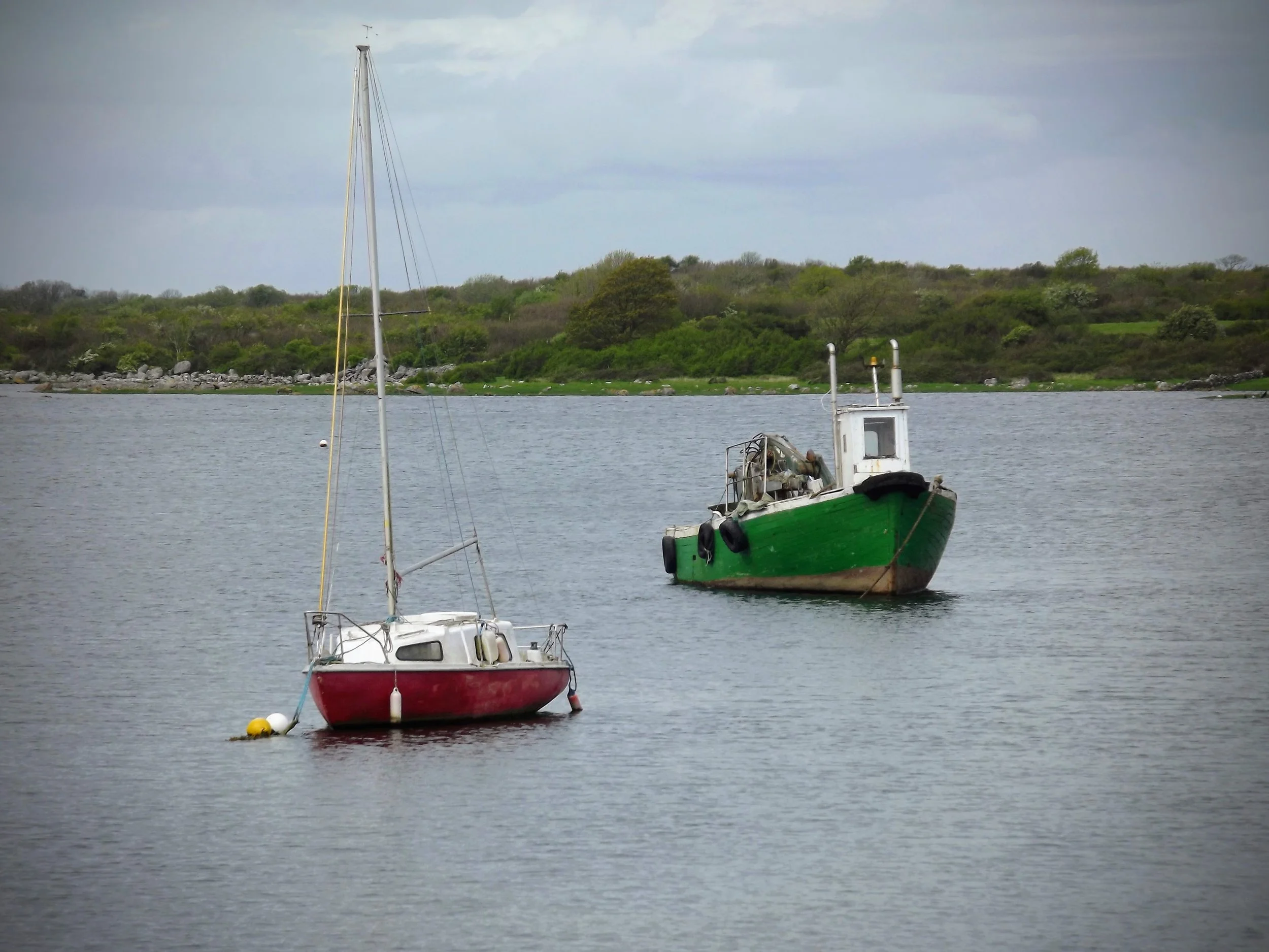 Two boats, a small sailboat and a motorboat, floating on calm water with a green, bush-covered shoreline in the background under a cloudy sky.