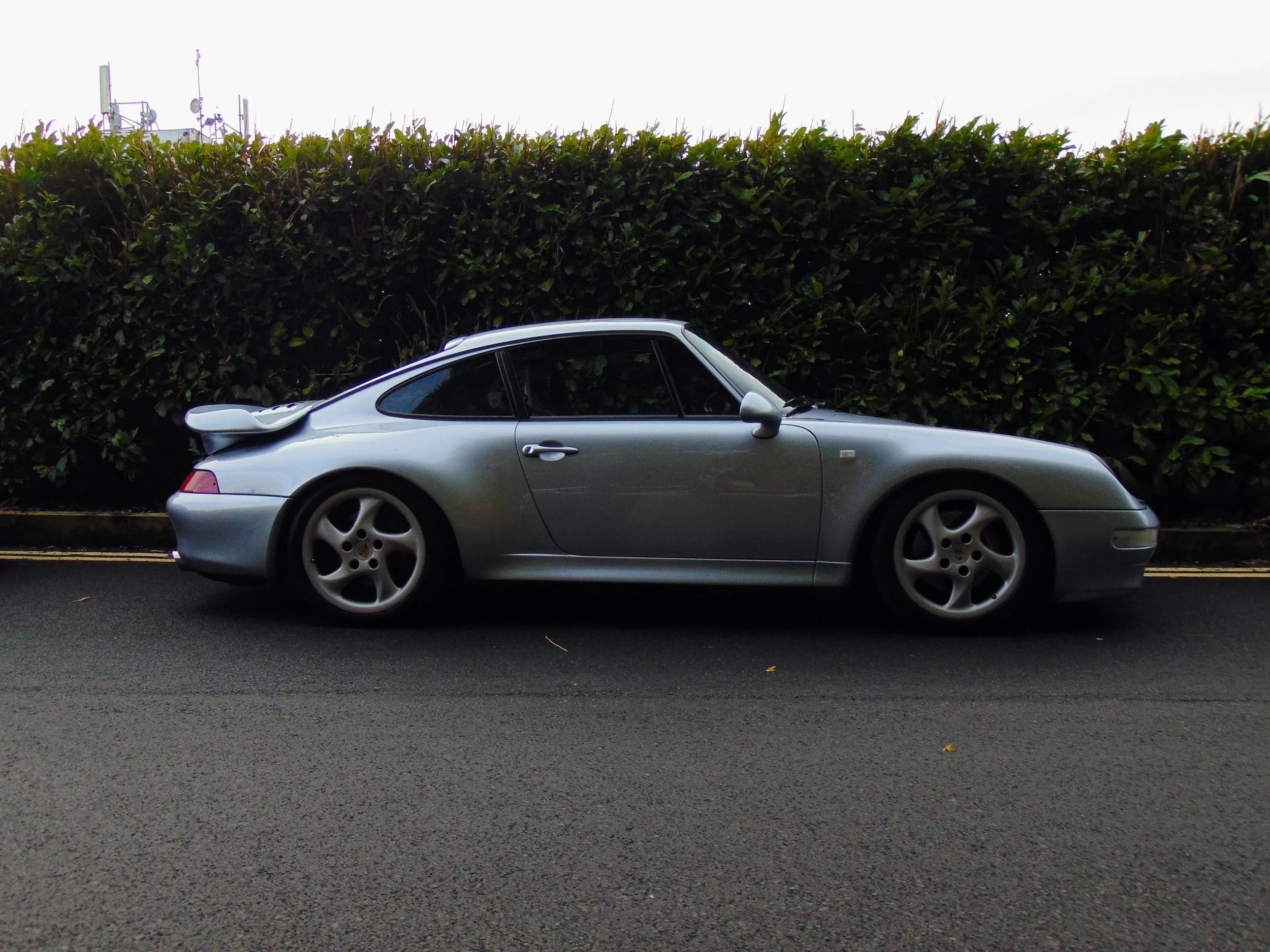 A silver Porsche 911 sports car parked on the side of a road with a hedge in the background.