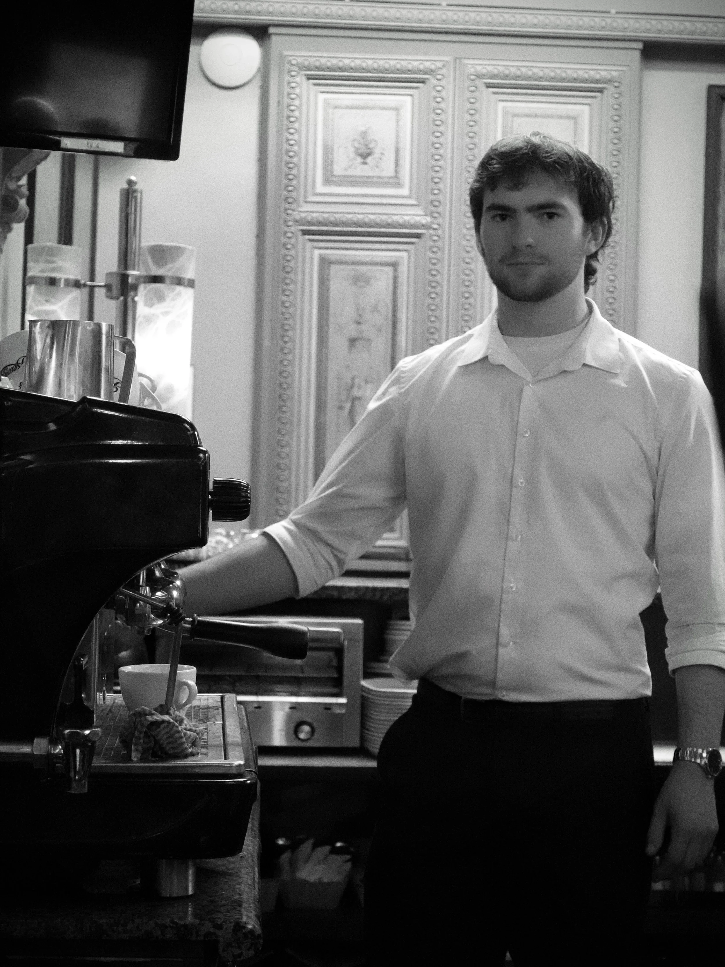 A man with dark hair and a beard, wearing a white shirt, standing in a restaurant or cafe setting, next to a coffee machine.