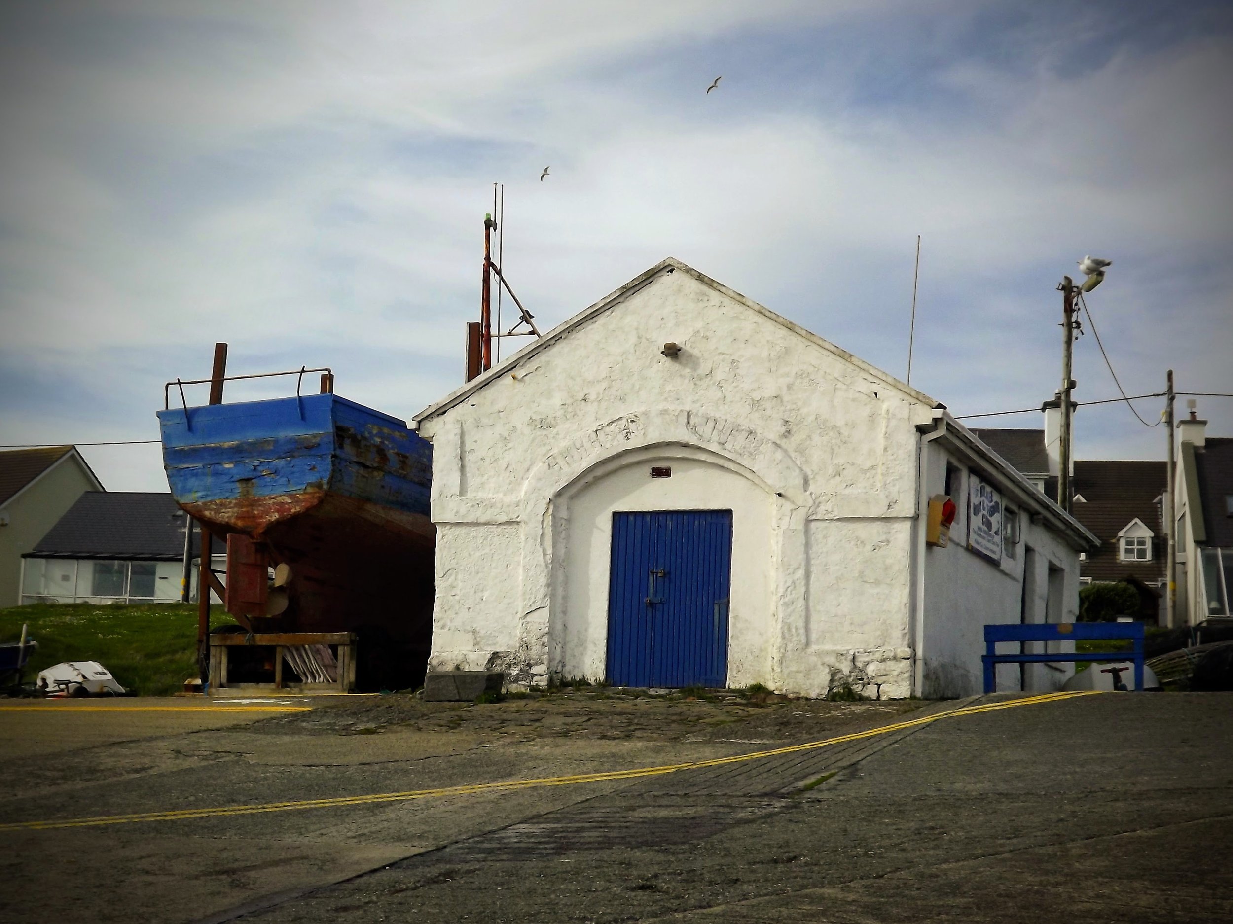 A small white building with a blue door and a blue boat on a stand next to it, with seagulls flying in the sky.