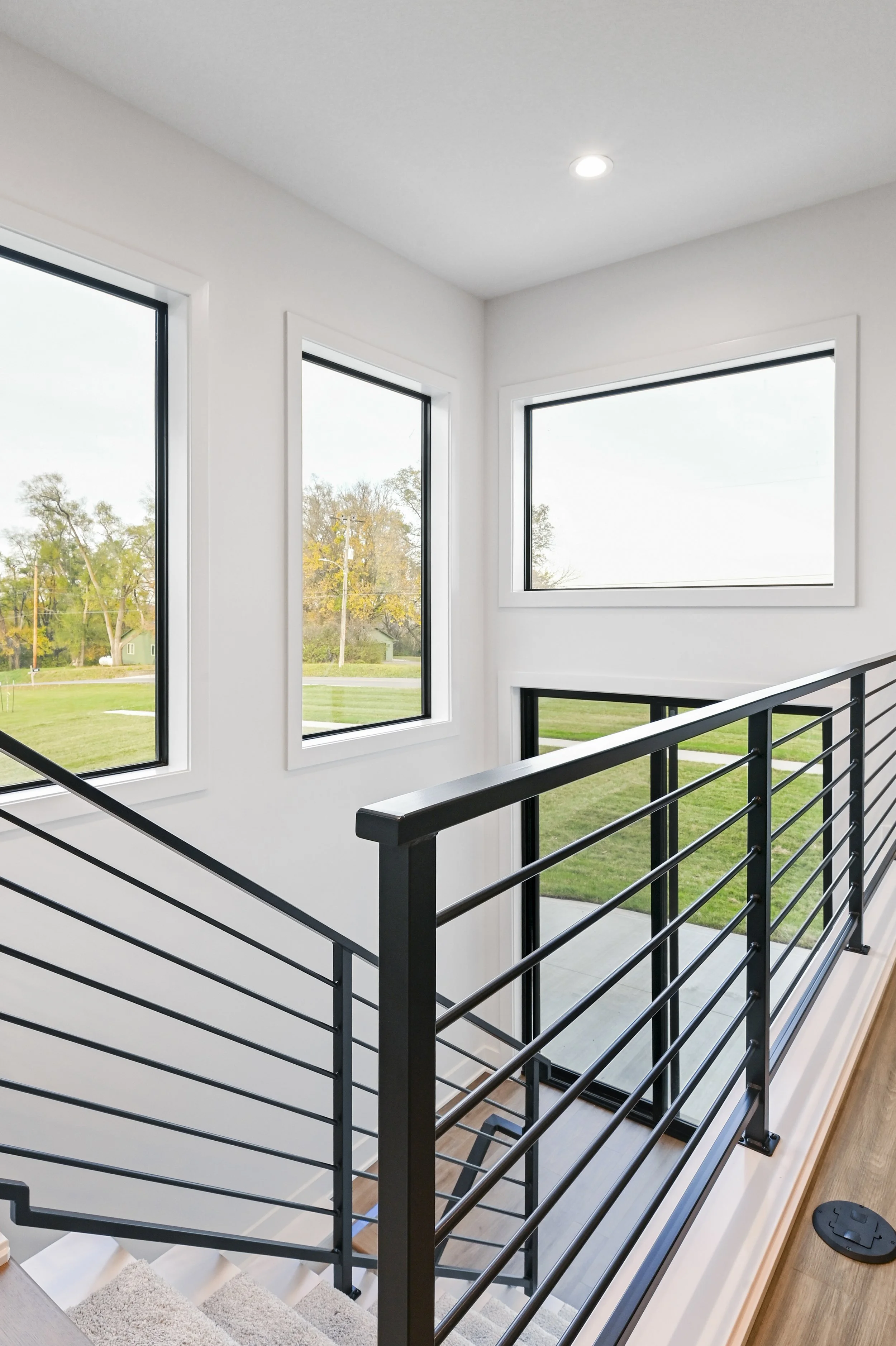 Interior view of a modern staircase with black metal railing and large windows allowing natural light, overlooking a green outdoor landscape.